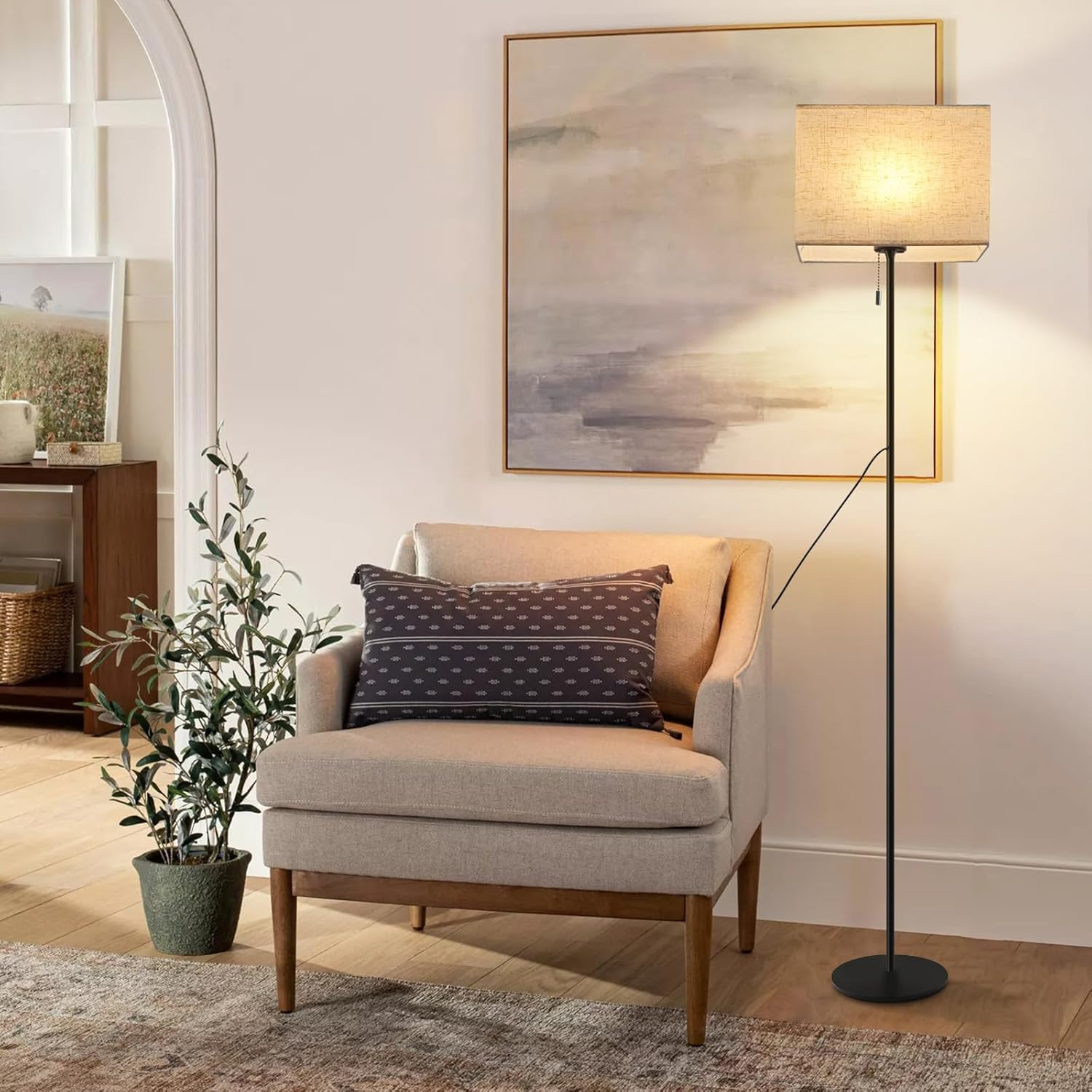 Wnętrze z lampą podłogową, fotelem, obrazem i rośliną, w odcieniach beżu i bieli.