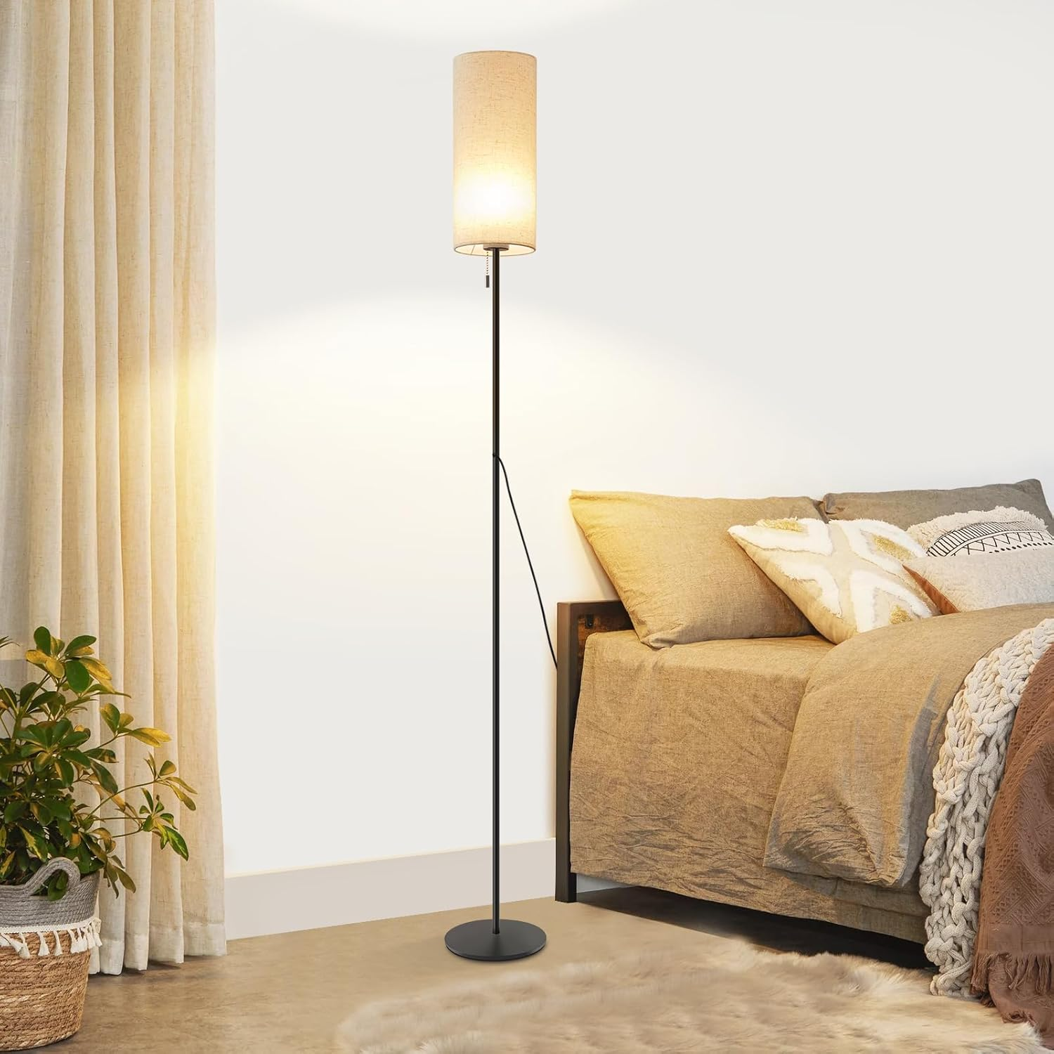 Wysoka lampa podłogowa z beżowym abażurem obok łóżka z poduszkami i rośliną.