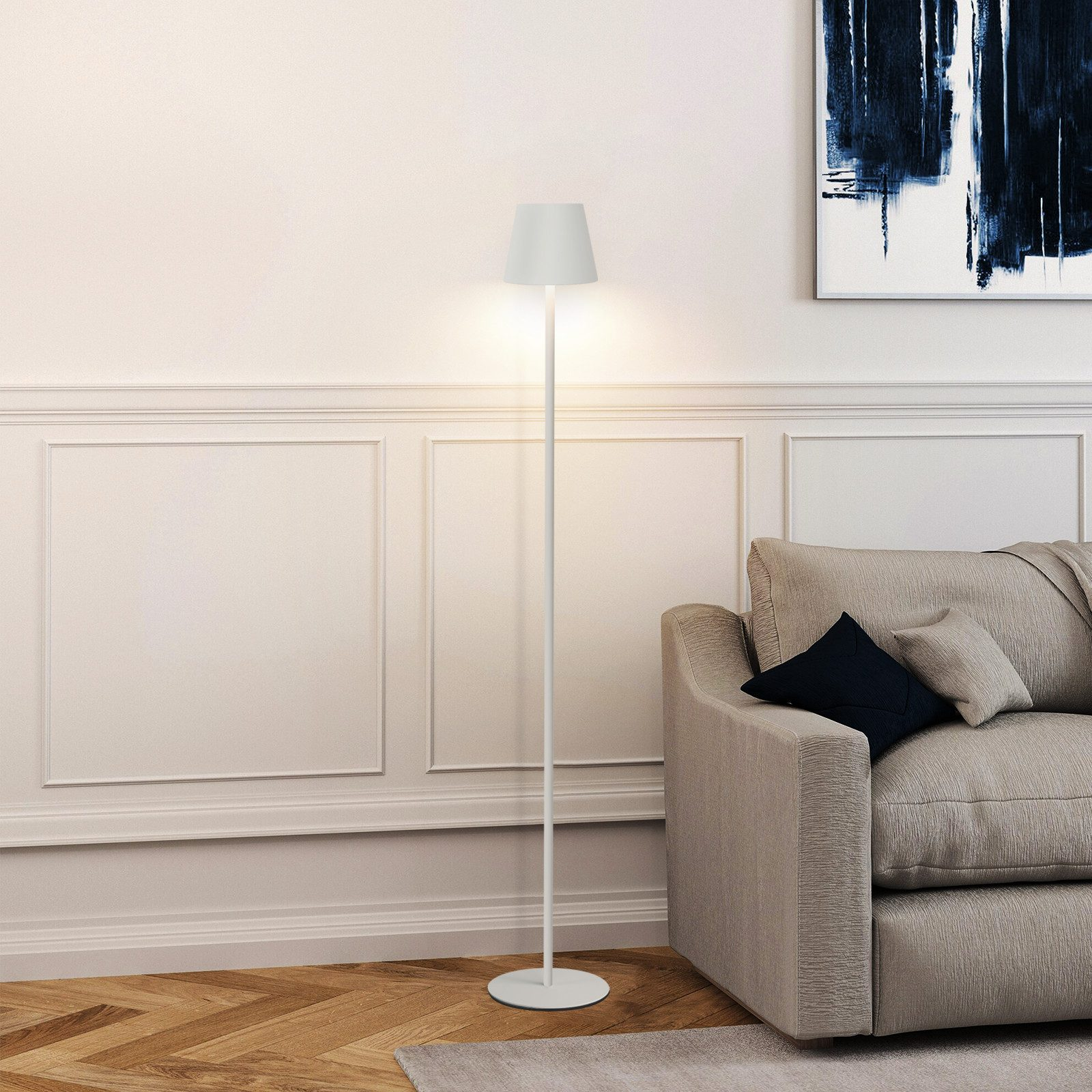 Lampada da terra bianca accanto a un divano e un dipinto. Illuminata. Muri bianchi e pavimento in legno.