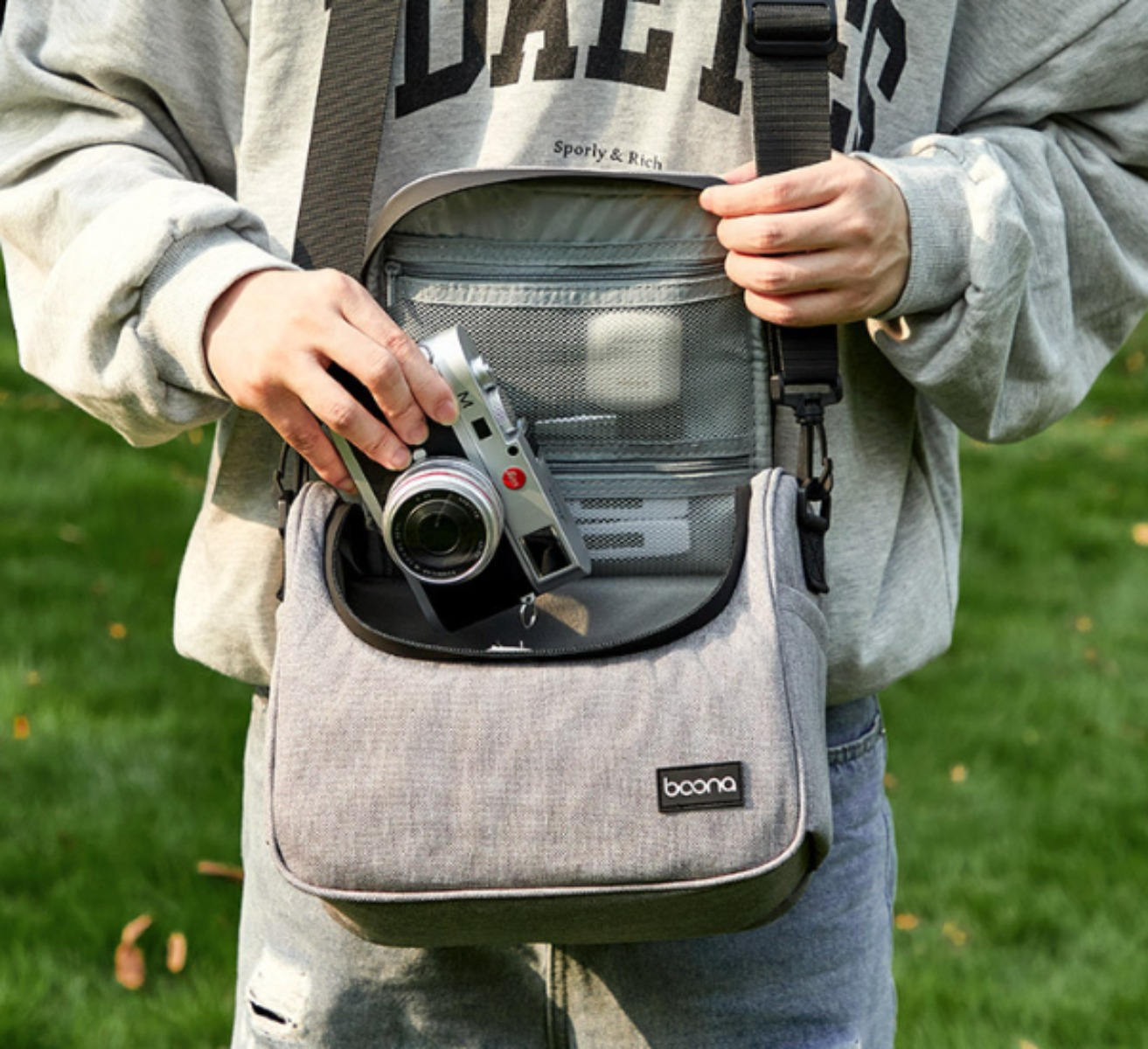 Person hält silberne Kamera in grauer Tasche, draußen mit grünem Gras.