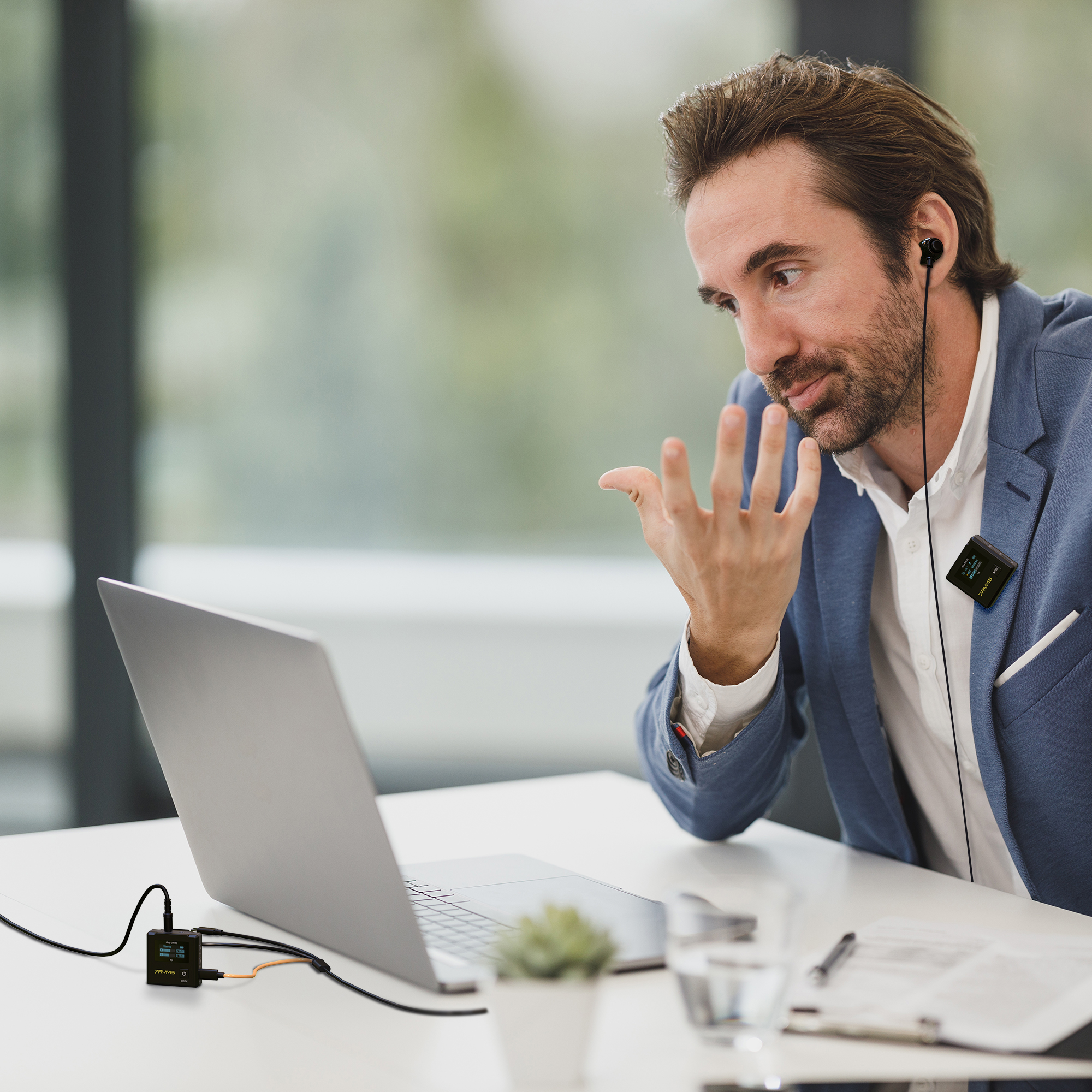 A man with a mic is talking while looking at a laptop. A glass and mic are visible.