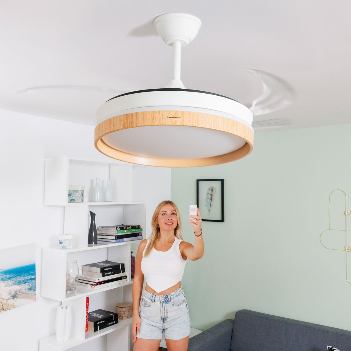 Mujer con mando a distancia controla ventilador de techo. Blanco con ribete efecto madera. Salón moderno.