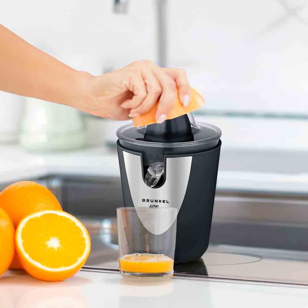 Una persona exprime media naranja en una exprimidora. Naranjas y un vaso de zumo están en la encimera.