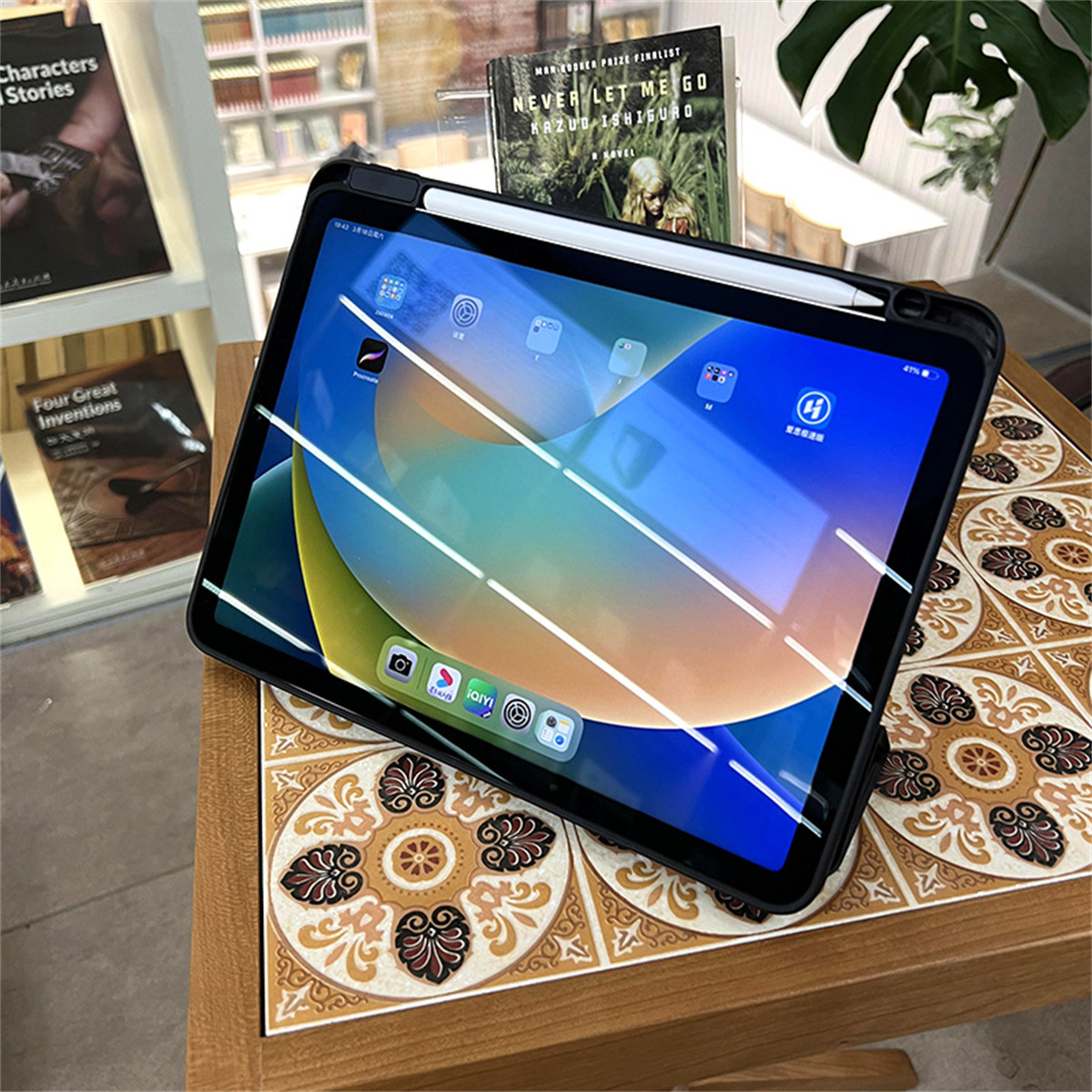 Ein Tablet mit Stift und Hülle liegt auf einem Holztisch, im Hintergrund Bücher und Regale.