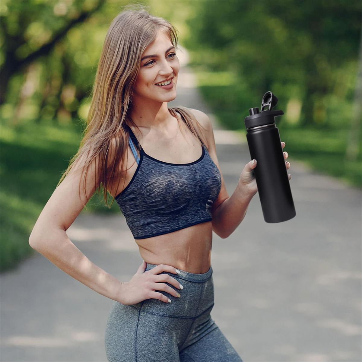 Frau in Sport-BH, hält schwarze Wasserflasche im Park, lächelt.