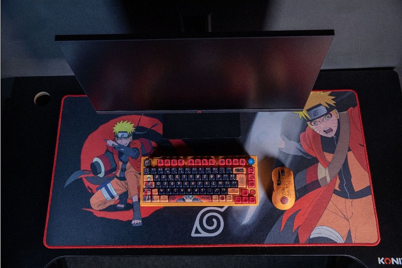 Configuration sur le thème de Naruto avec un moniteur, un tapis de souris Naruto, un clavier et une souris sur un bureau noir.