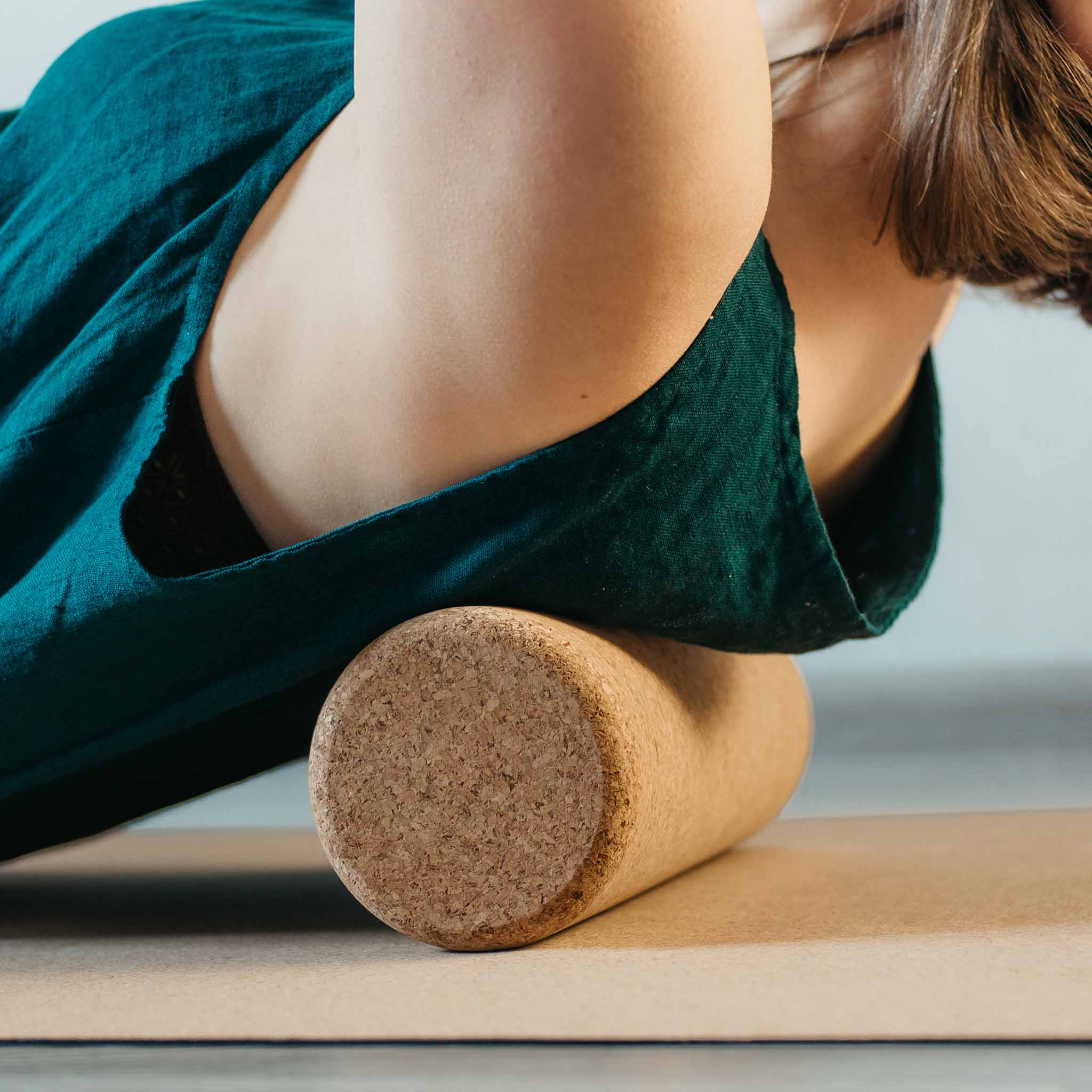 Person in tealfarbenem Oberteil verwendet eine Korkrolle auf einer Yogamatte. Die Person liegt auf dem Rücken.