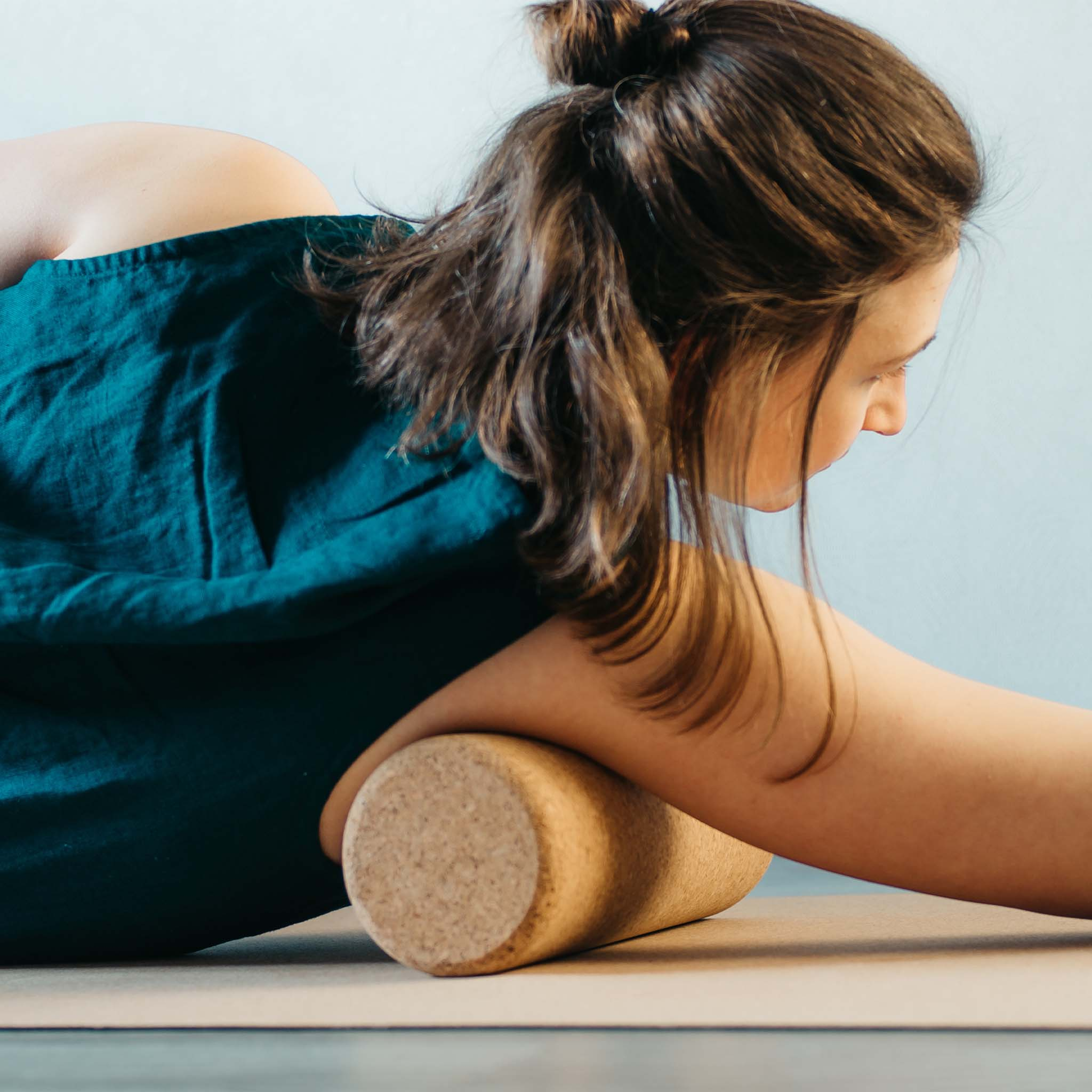 Frau auf Yogamatte mit Korkrolle. Sie trägt ein türkisfarbenes Hemd, Fokus auf ihrem Rücken.