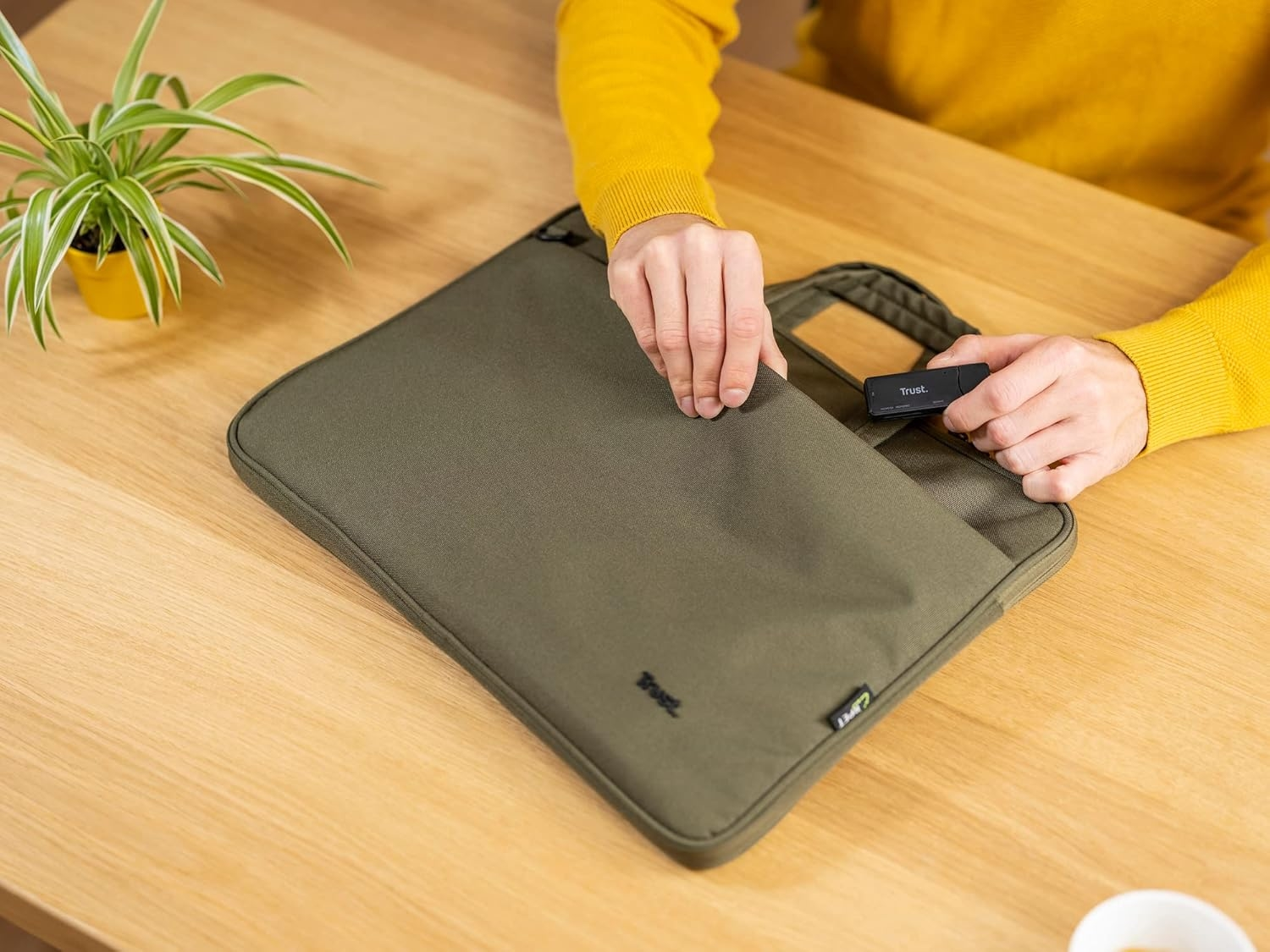 Osoba wkładająca czytnik kart Trust do zielonej torby na laptopa na drewnianym stole. Żółta koszula.