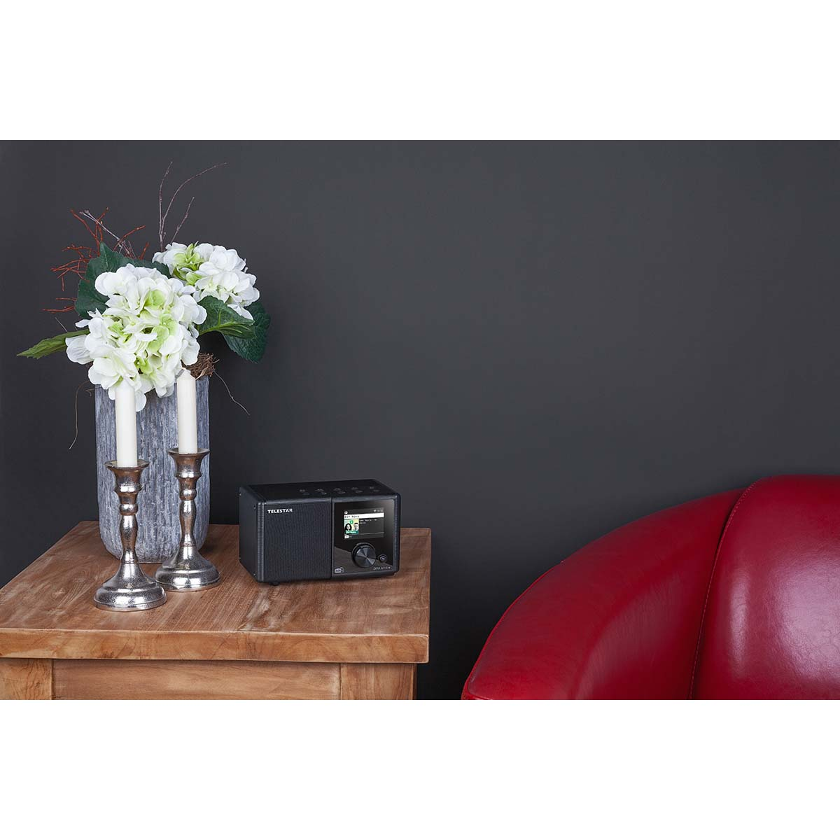 Ein schwarzes Radio auf einem Holztisch, mit Kerzen, Blumen und einem roten Sessel im Hintergrund.