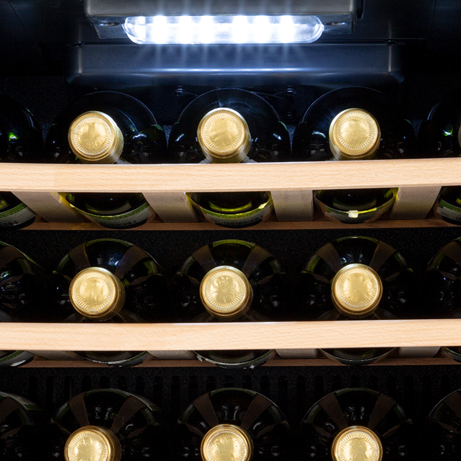 Botellas de vino en estantes dentro de un refrigerador. Hay una luz en la parte superior. Estantes de madera.