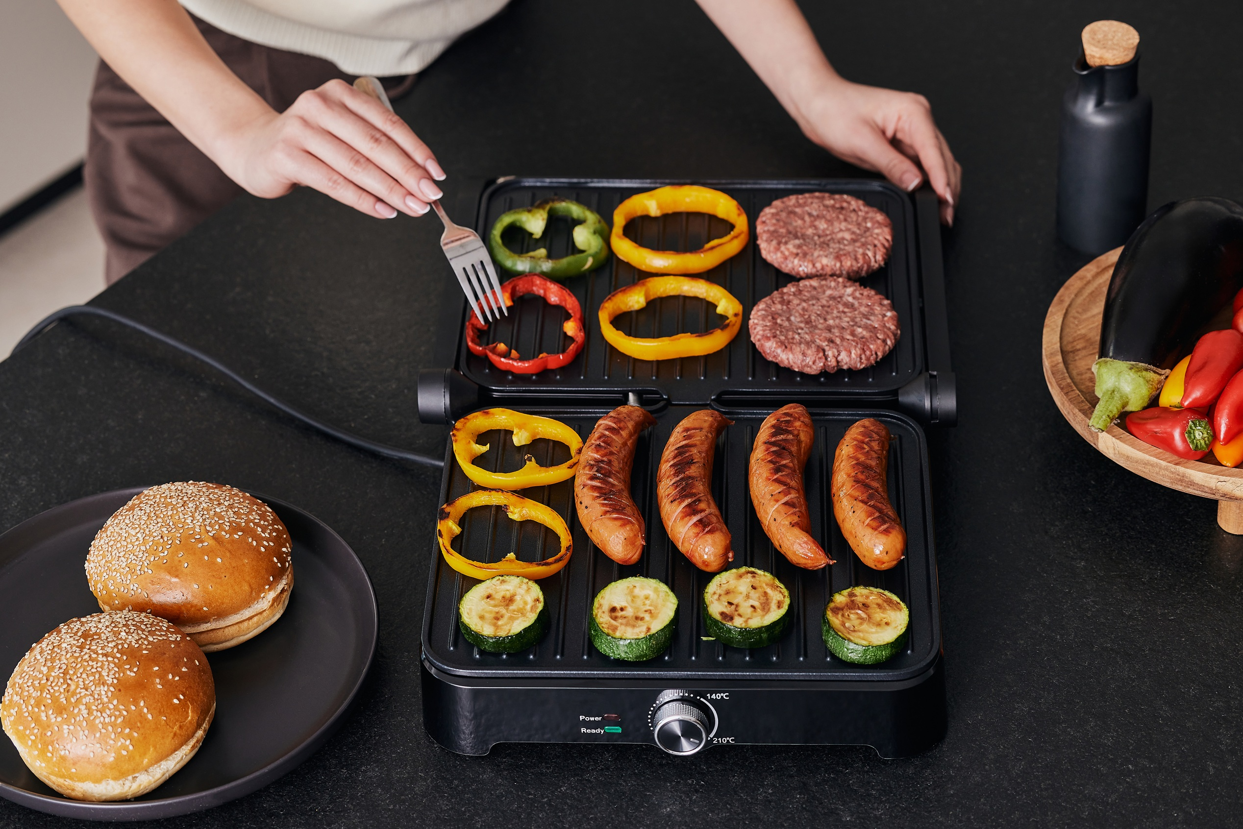 Hände grillen Essen auf einem schwarzen Küchentischgrill: Burger, Würstchen, Paprika und Zucchini.
