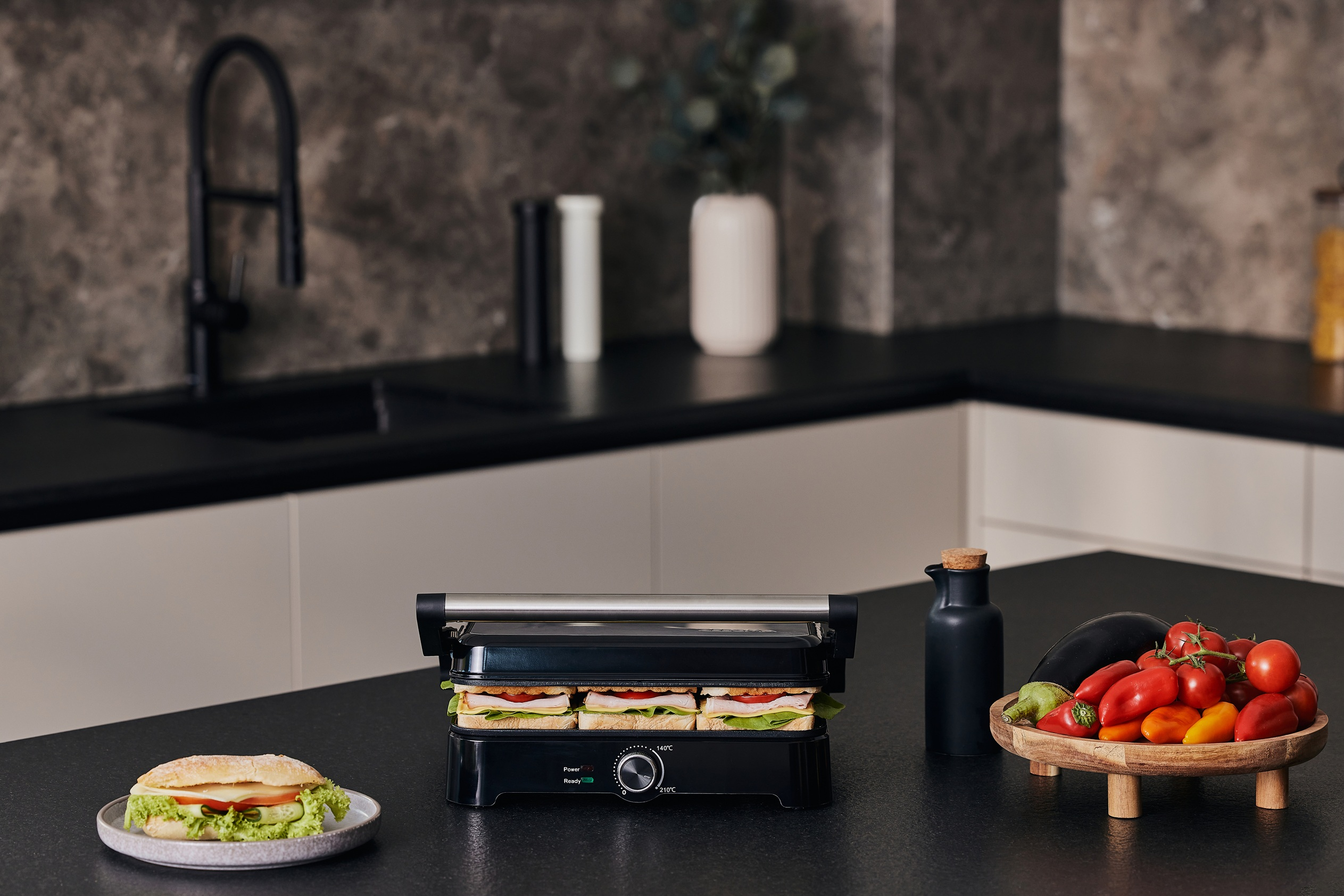 Sandwichmaker mit Sandwiches im Inneren. Ein Teller mit Sandwich links, Gemüse.