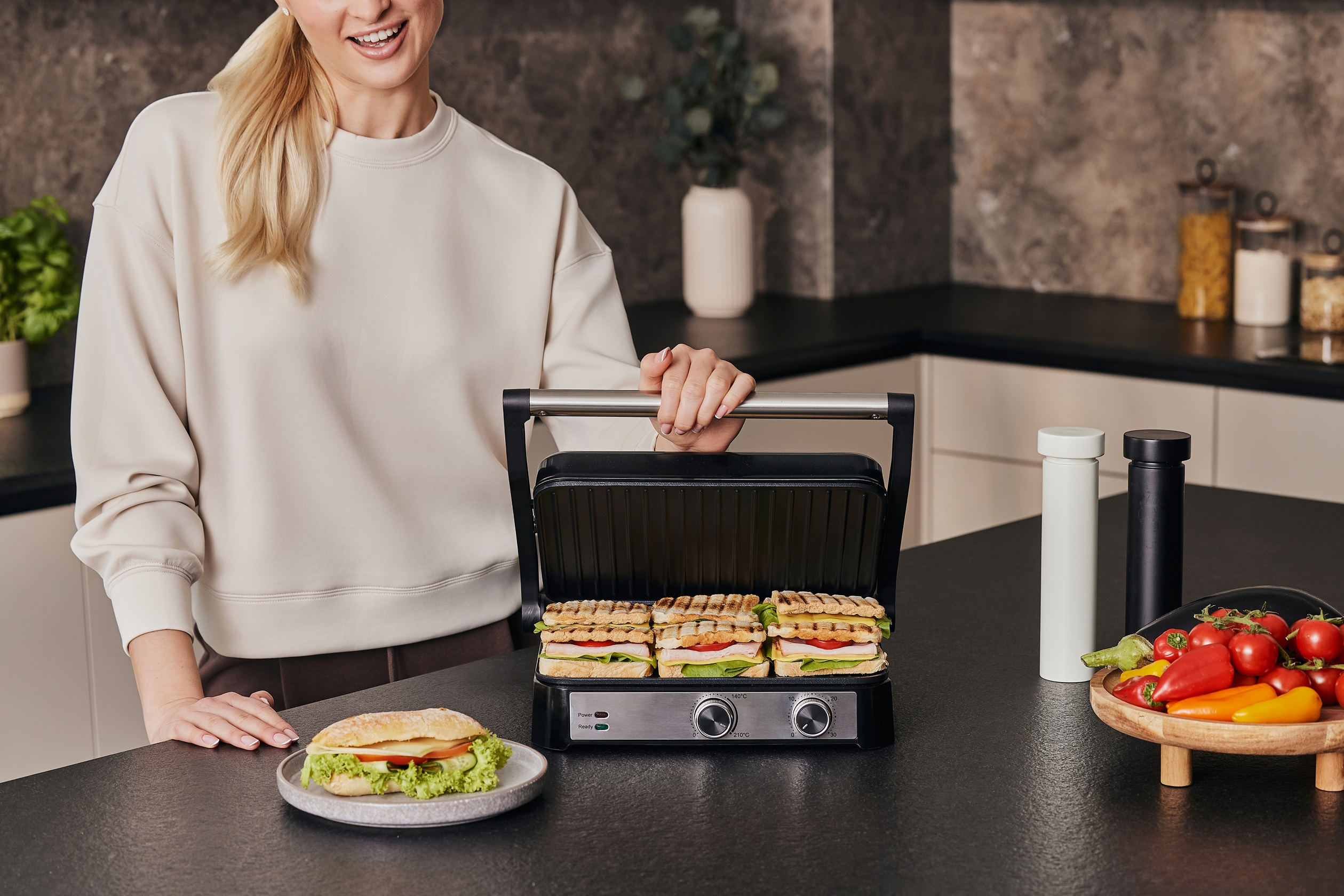 Frau mit Sandwich auf einem Teller und Sandwichgrill mit Sandwiches. Gemüse ist sichtbar.