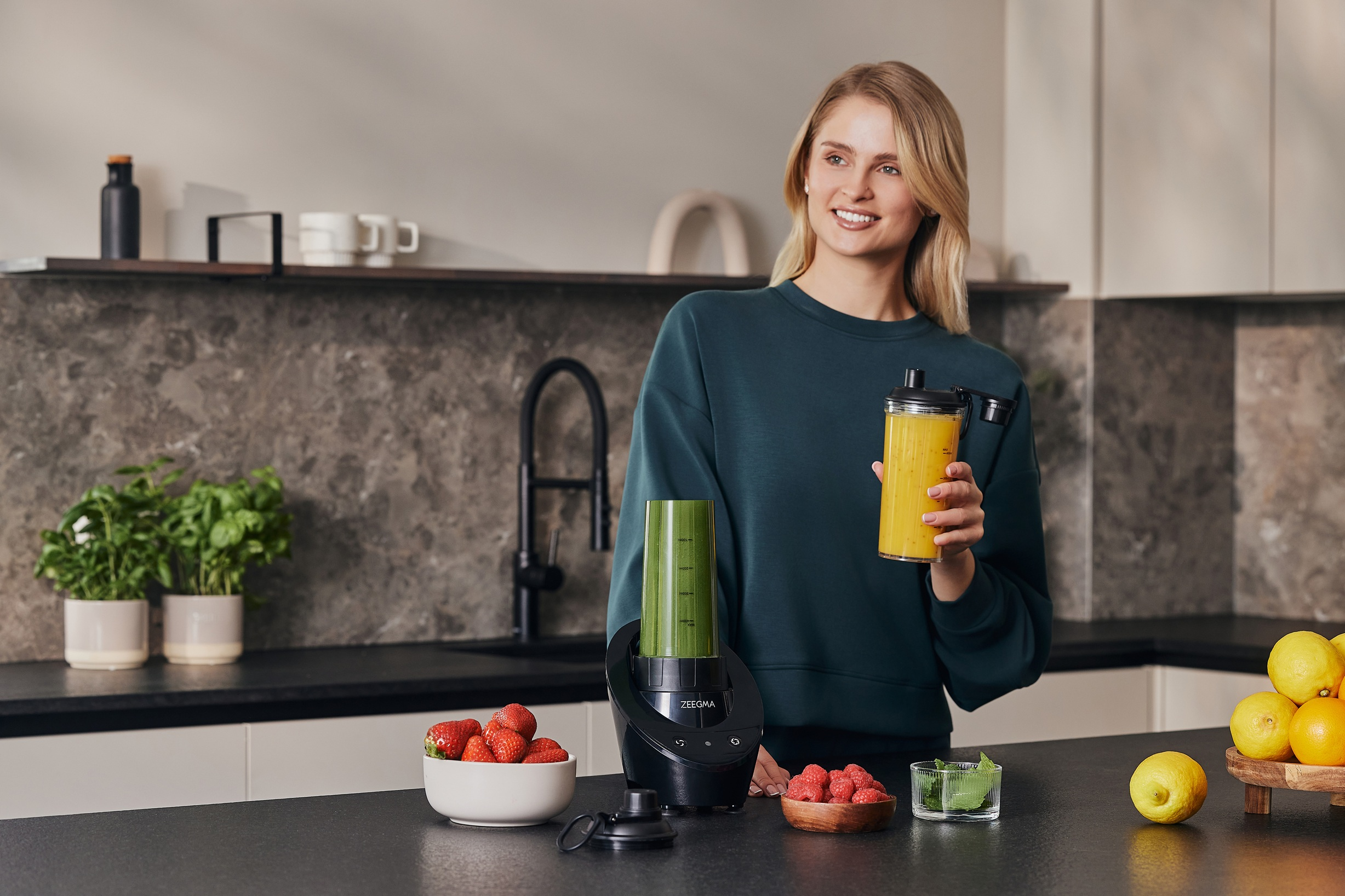 Frau hält Smoothie, Mixer und Zutaten auf der Theke. Küchenhintergrund.