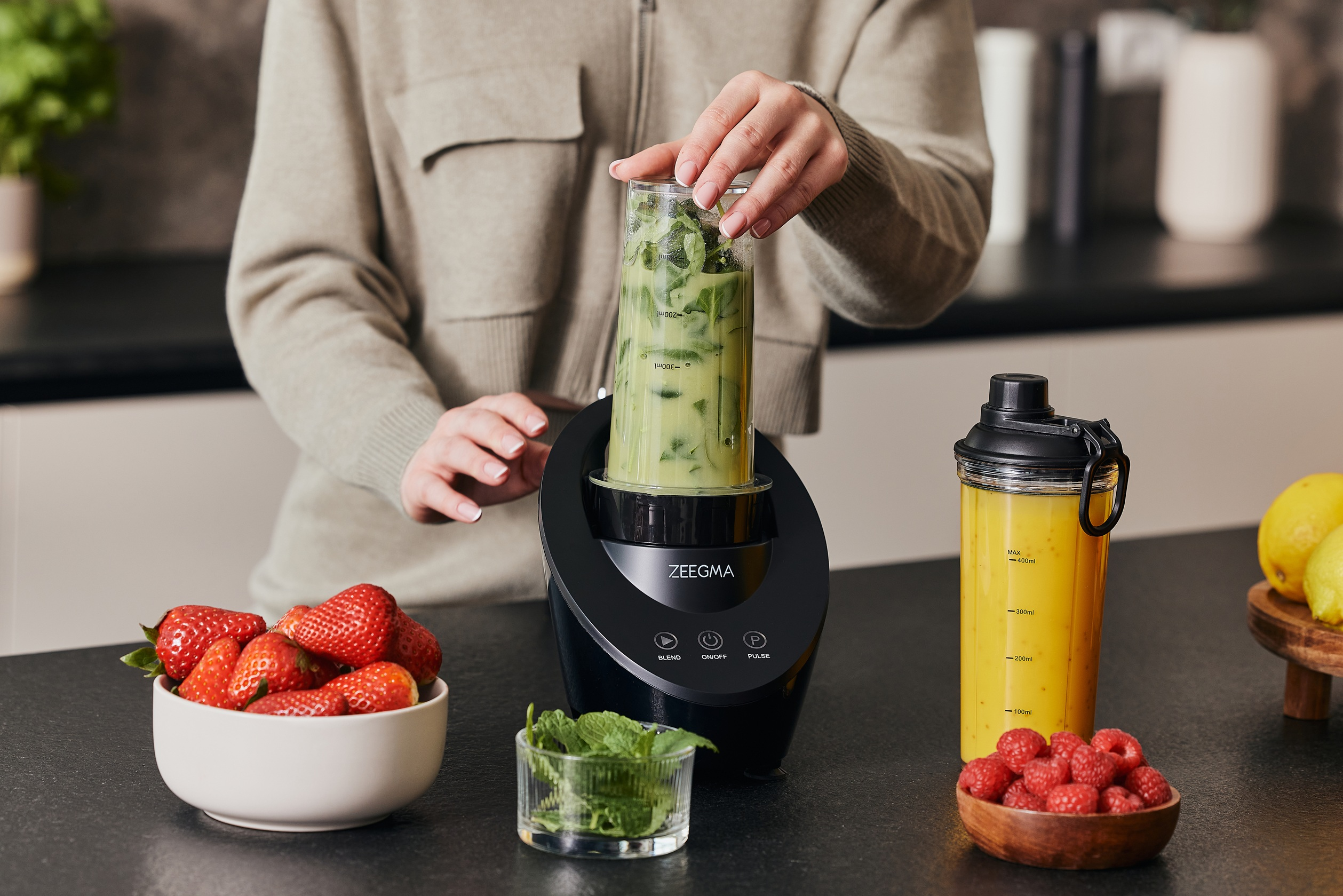 Frau, die einen Mixerbecher mit grüner Flüssigkeit in einen schwarzen Mixer stellt. Erdbeeren in der Nähe.