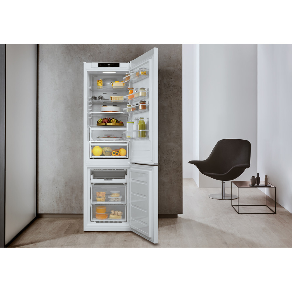 Refrigerador blanco con puertas abiertas, mostrando estantes con comida. Silla negra y mesa pequeña al fondo.