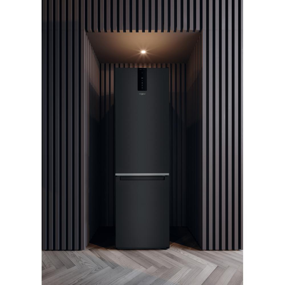 Un refrigerador negro en un entorno oscuro. Es alto con congelador inferior y puerta cerrada.