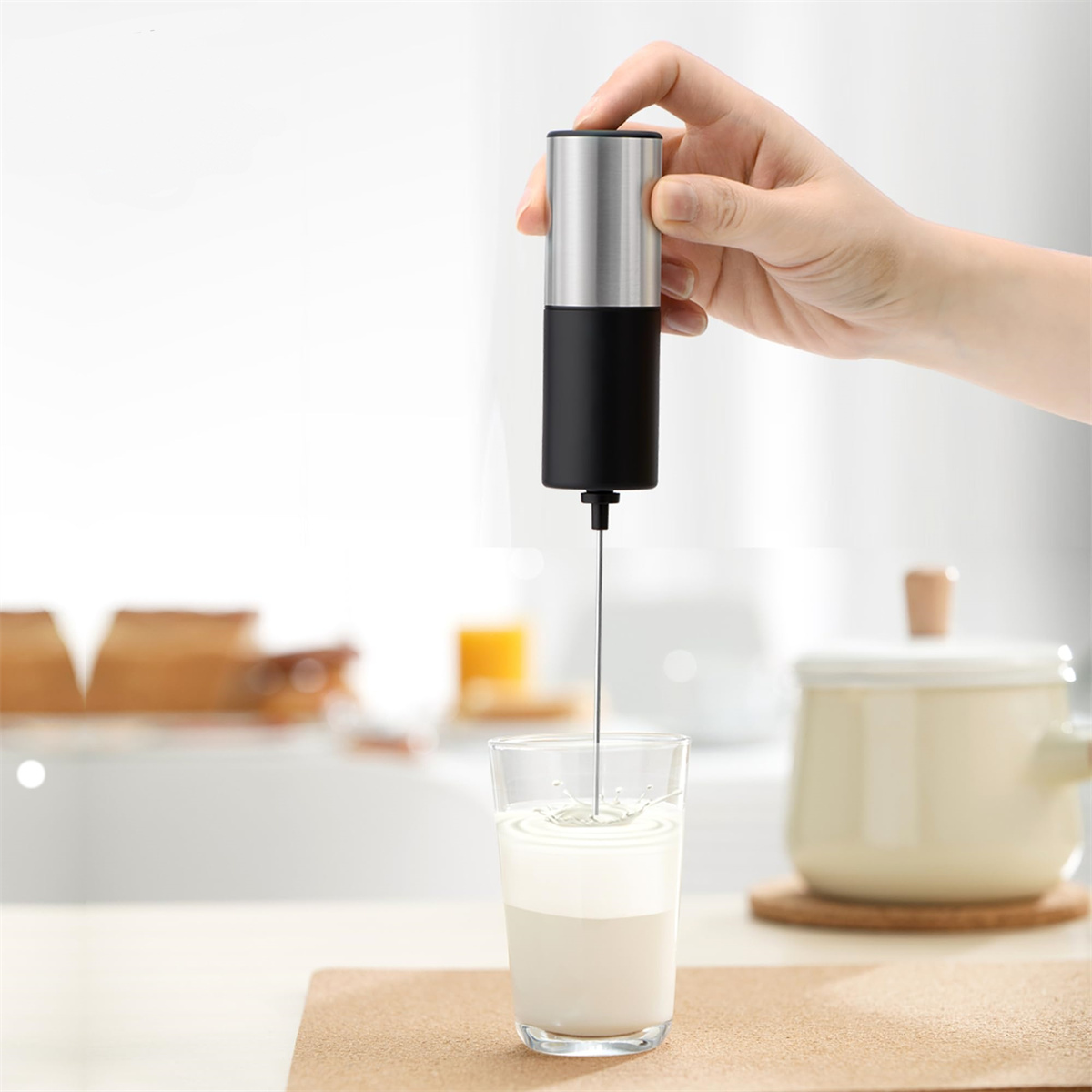 Eine Hand, die einen Milchaufschäumer über einem Glas Milch hält und Schaum erzeugt. Der Hintergrund ist eine Küchenumgebung.