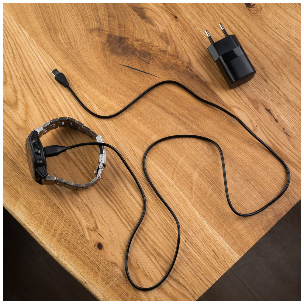 Eine Smartwatch, die auf einem Holztisch geladen wird. Ladegerät und Kabel sind sichtbar. Schwarzes Ladegerät, Holztisch.