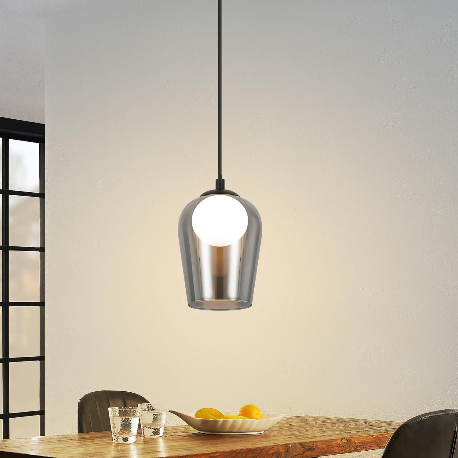 Lampa wisząca z szarym szklanym kloszem i białą żarówką w jadalni. Jest stół i szklanki.