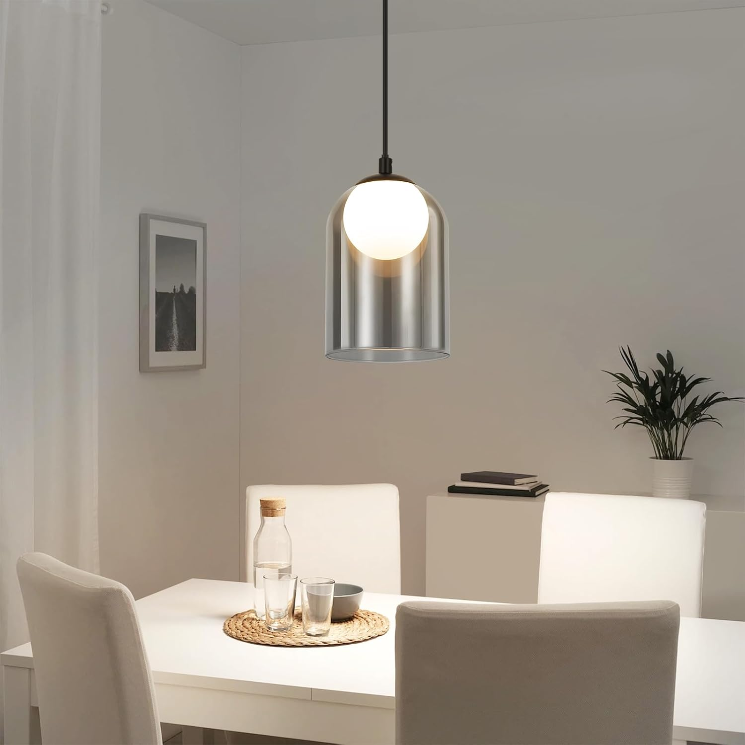 Lampa wisząca nad białym stołem. Lampa ma szklany klosz, a na stole stoi roślina.