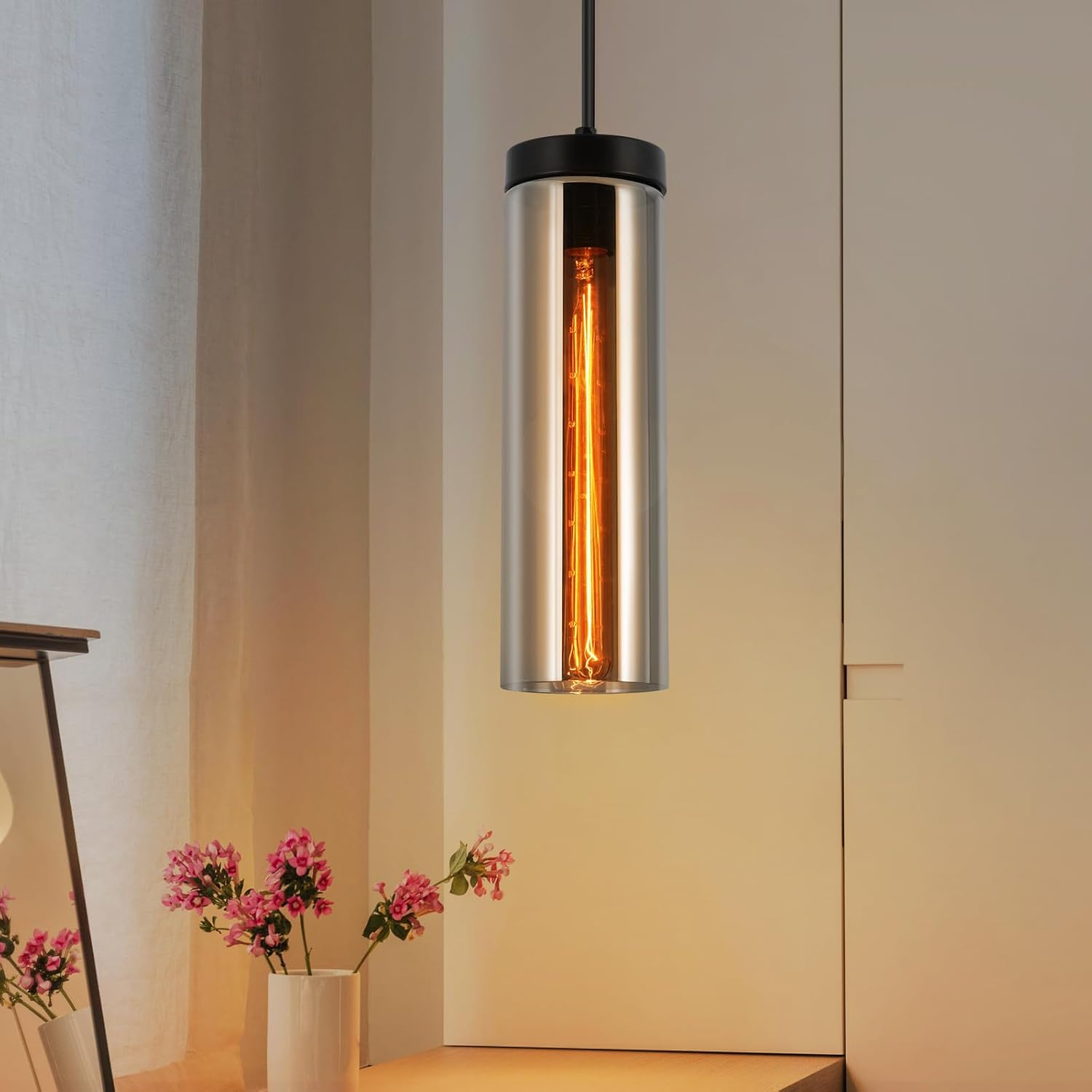 Eine einzelne Pendelleuchte mit zylindrischem Glasschirm, einer warmtonigen Glühbirne und schwarzen Armaturen.