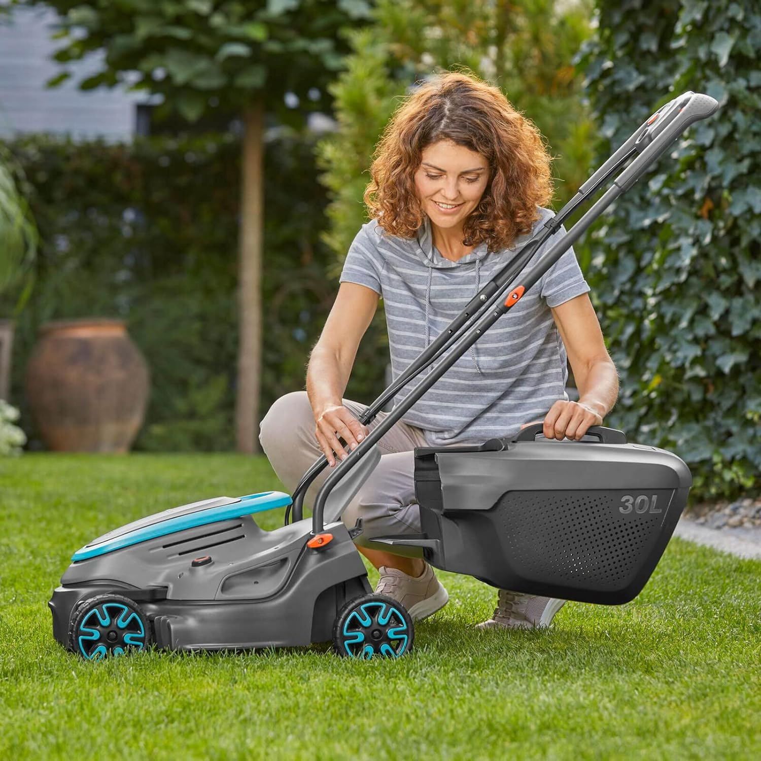 Frau bedient einen grau-blauen Rasenmäher in einer grünen, grasbewachsenen Fläche, Bäume im Hintergrund.