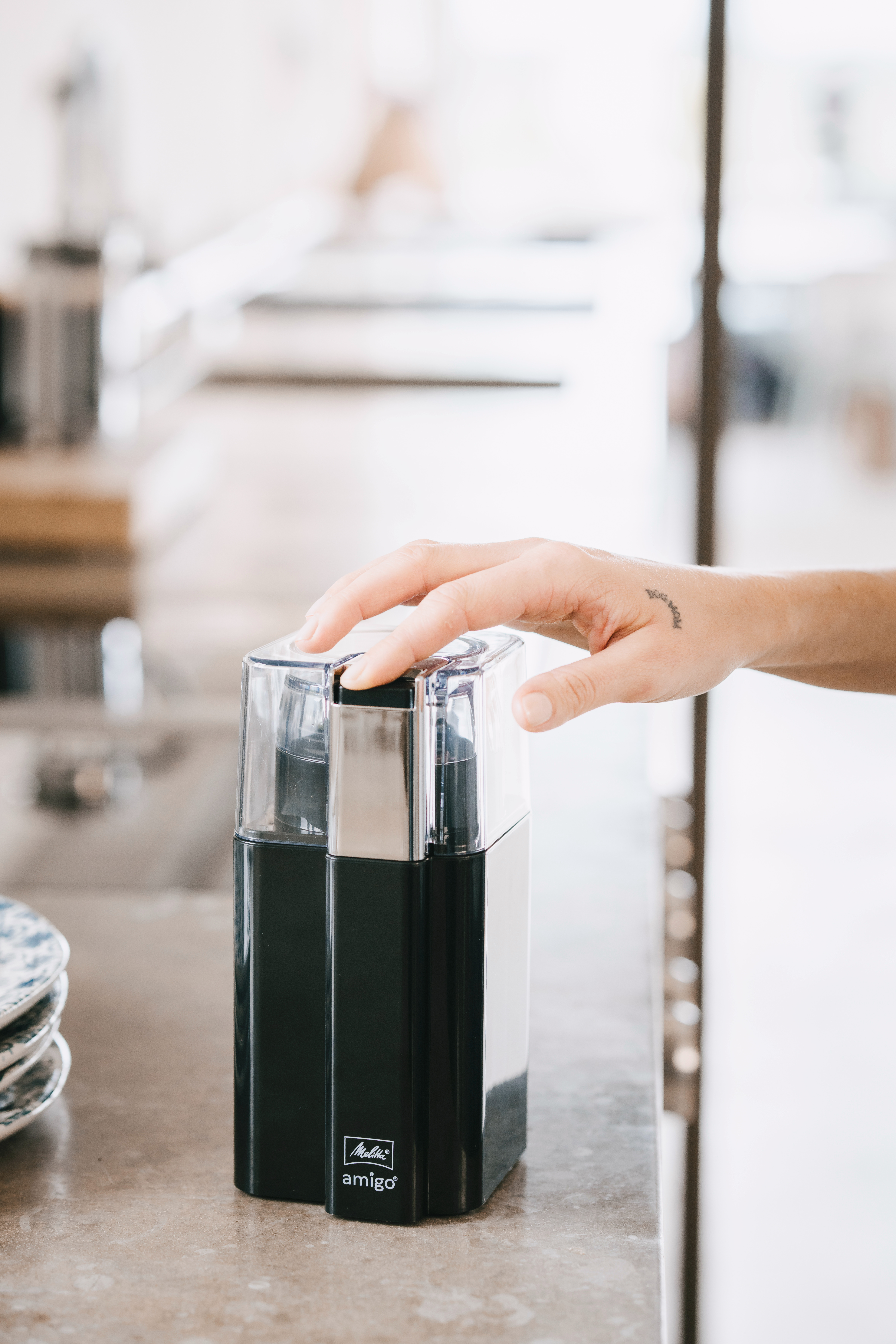 Ręka trzyma czarny młynek do kawy, obok talerza z naczyniami.