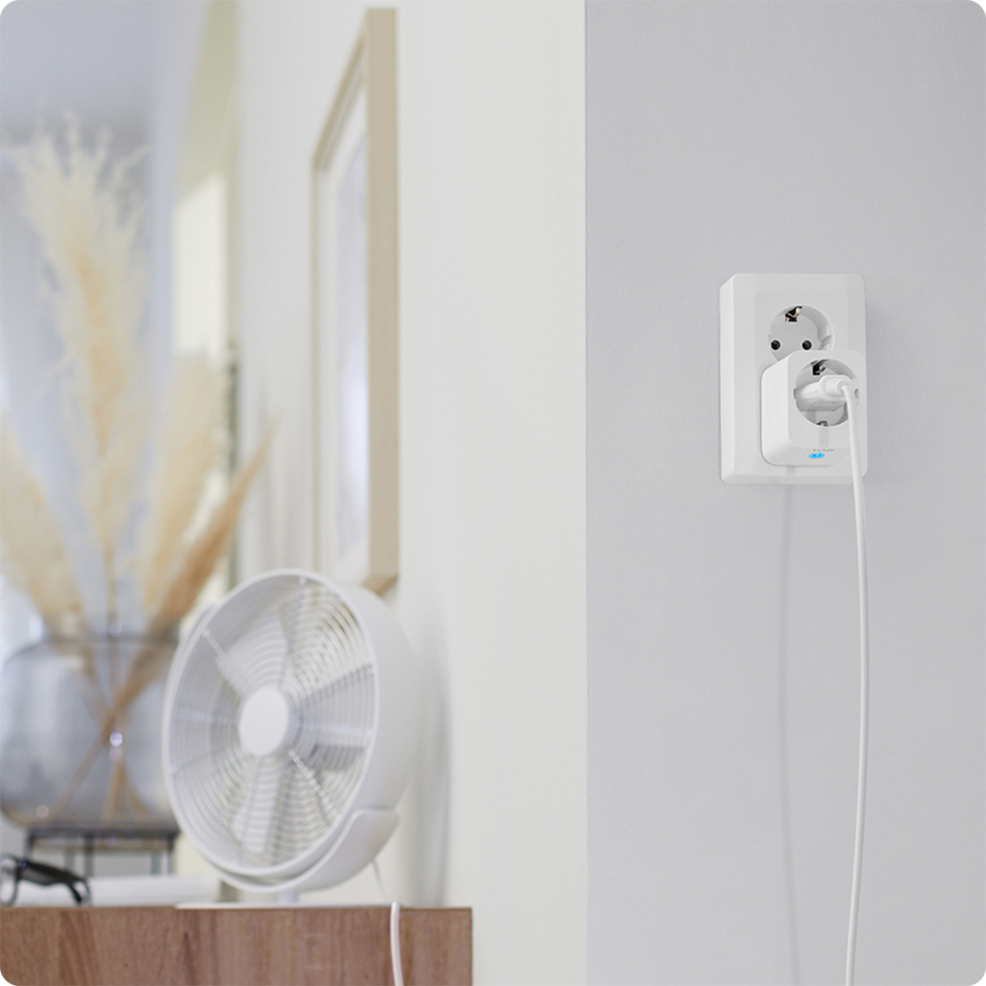 Een witte ventilator op een tafel, een stopcontact en een fotolijst zijn zichtbaar.