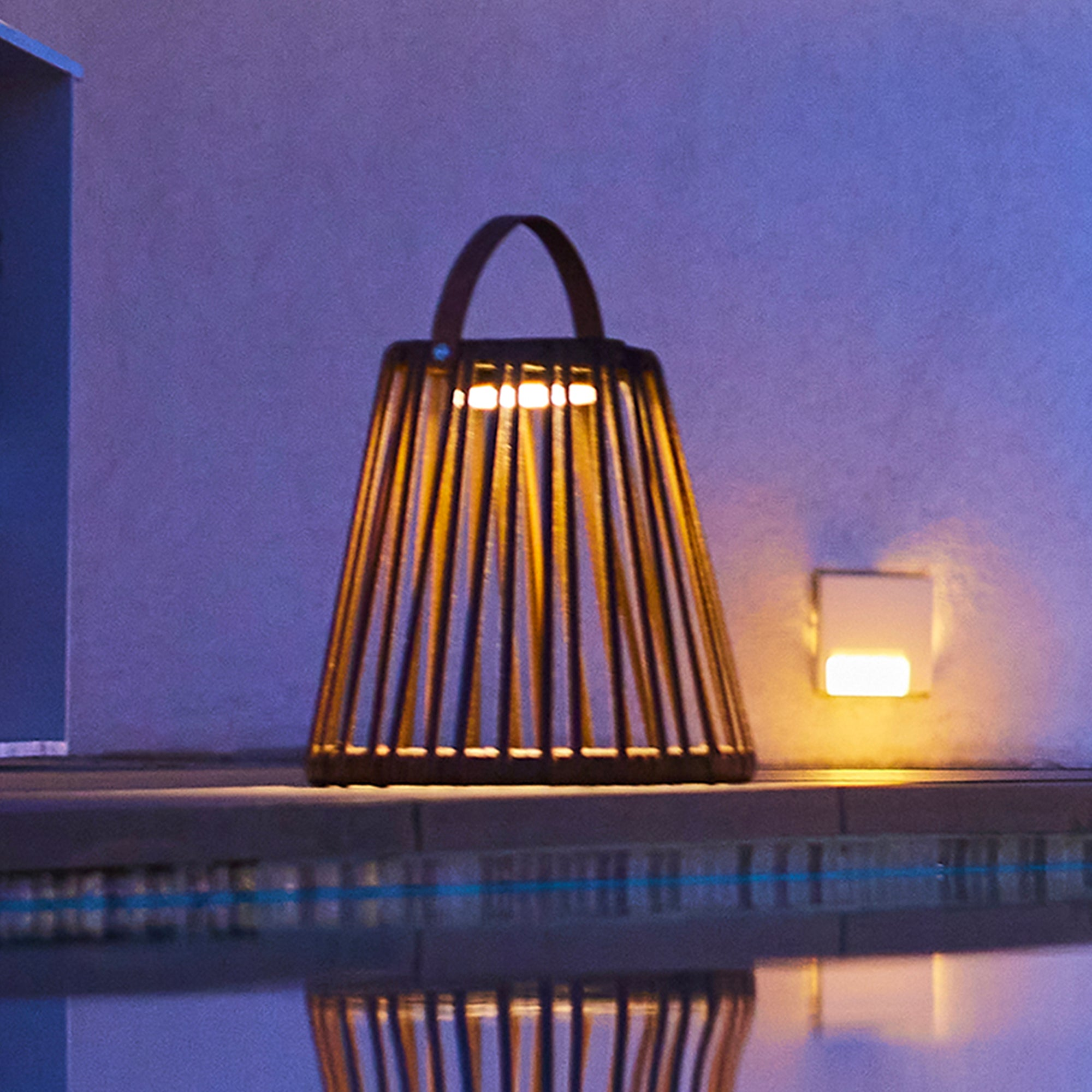 Eine braune Laterne mit Griff steht in der Nähe eines Pools, beleuchtet von einer Wandlampe und Nachtlichtern. Nachtszene.