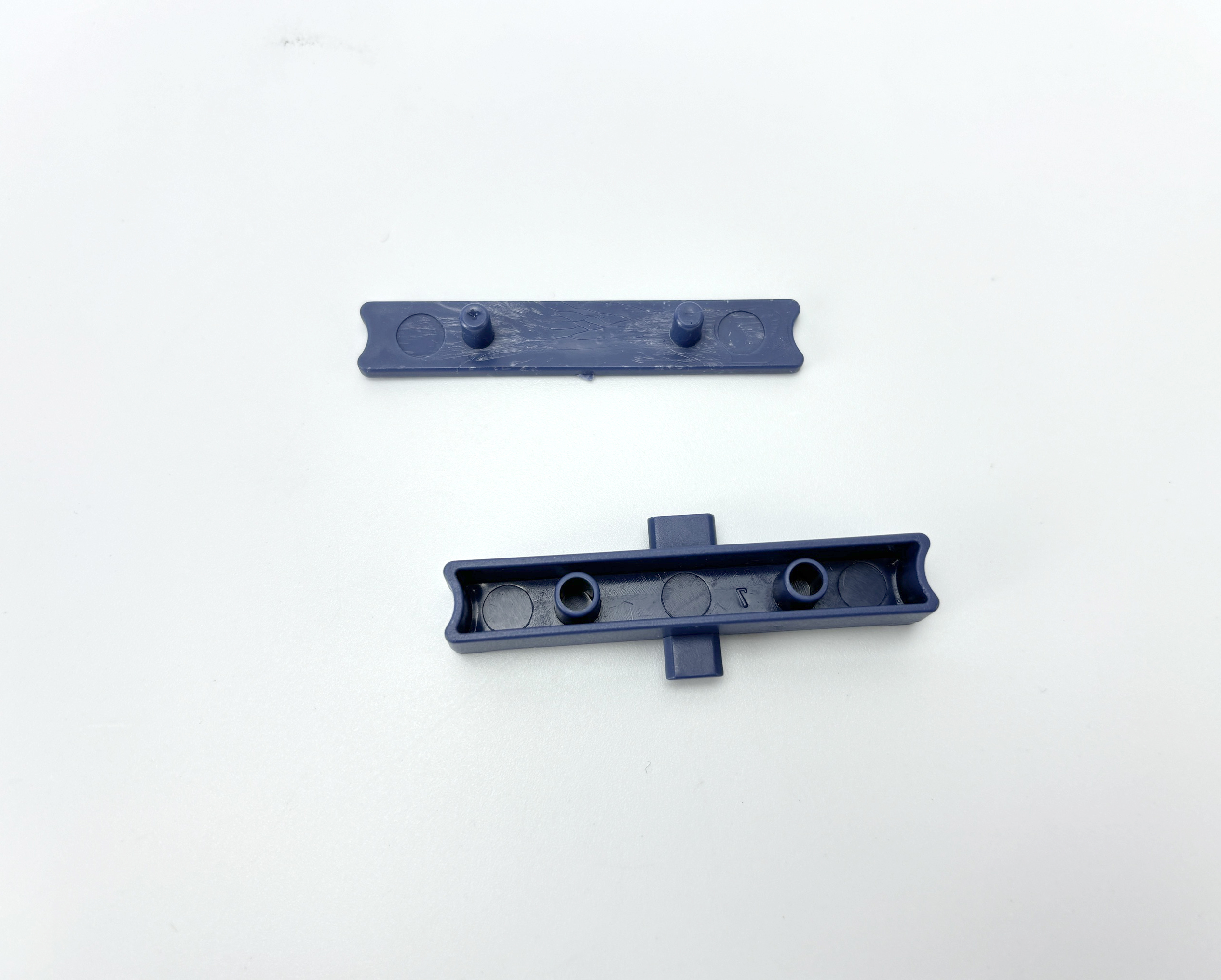 Two navy blue plastic pieces lie on a white surface. Each has holes and a unique shape.