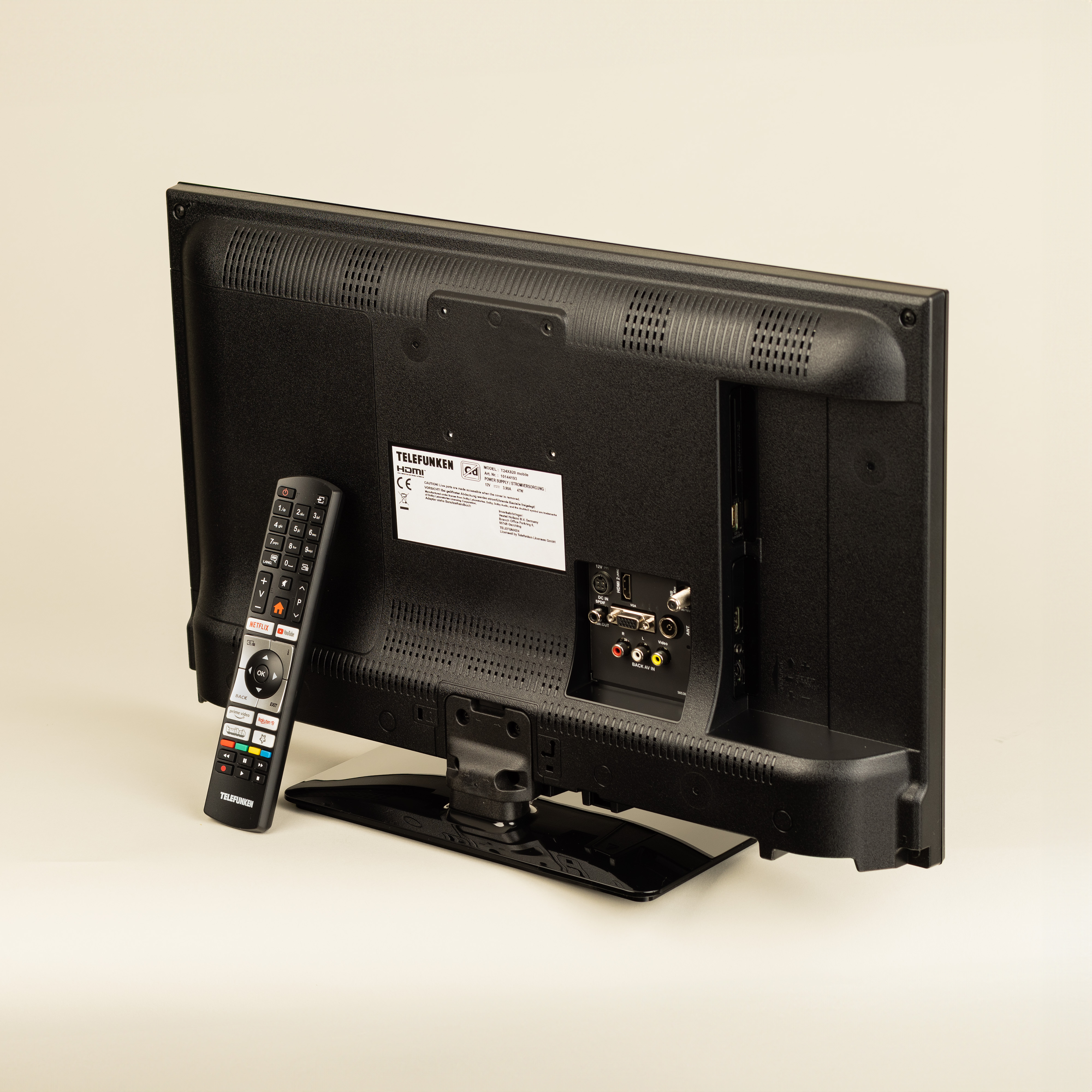 Die Rückseite eines schwarzen Fernsehers mit Fernbedienung. Es hat Anschlüsse und ein Etikett, vor einem hellcremefarbenen Hintergrund.