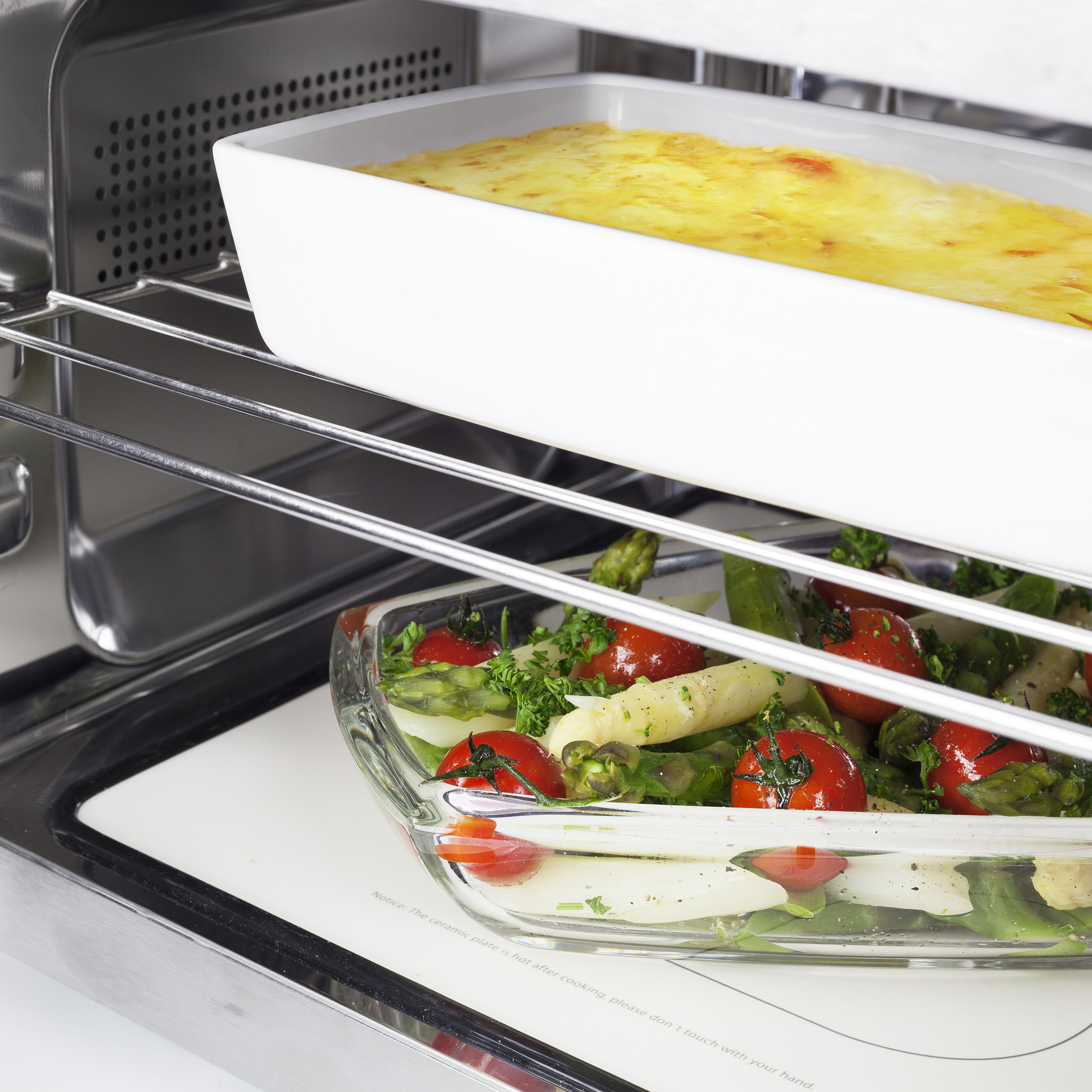 Un plat en verre avec des légumes cuits et un plat à gratin au-dessus d'un intérieur de four argenté.