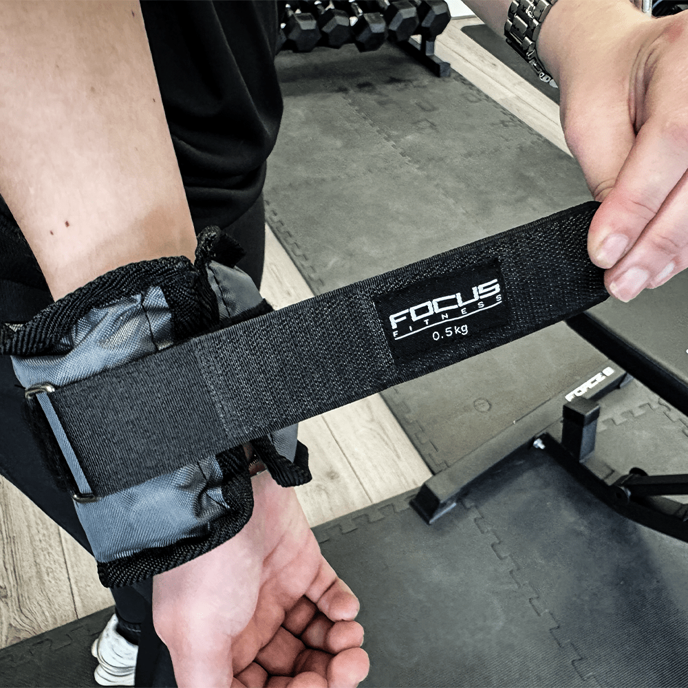 Persoon in een sportschool bevestigt een zwarte Focus Fitness polsgewicht. Het gewicht is gelabeld 0,5 kg.
