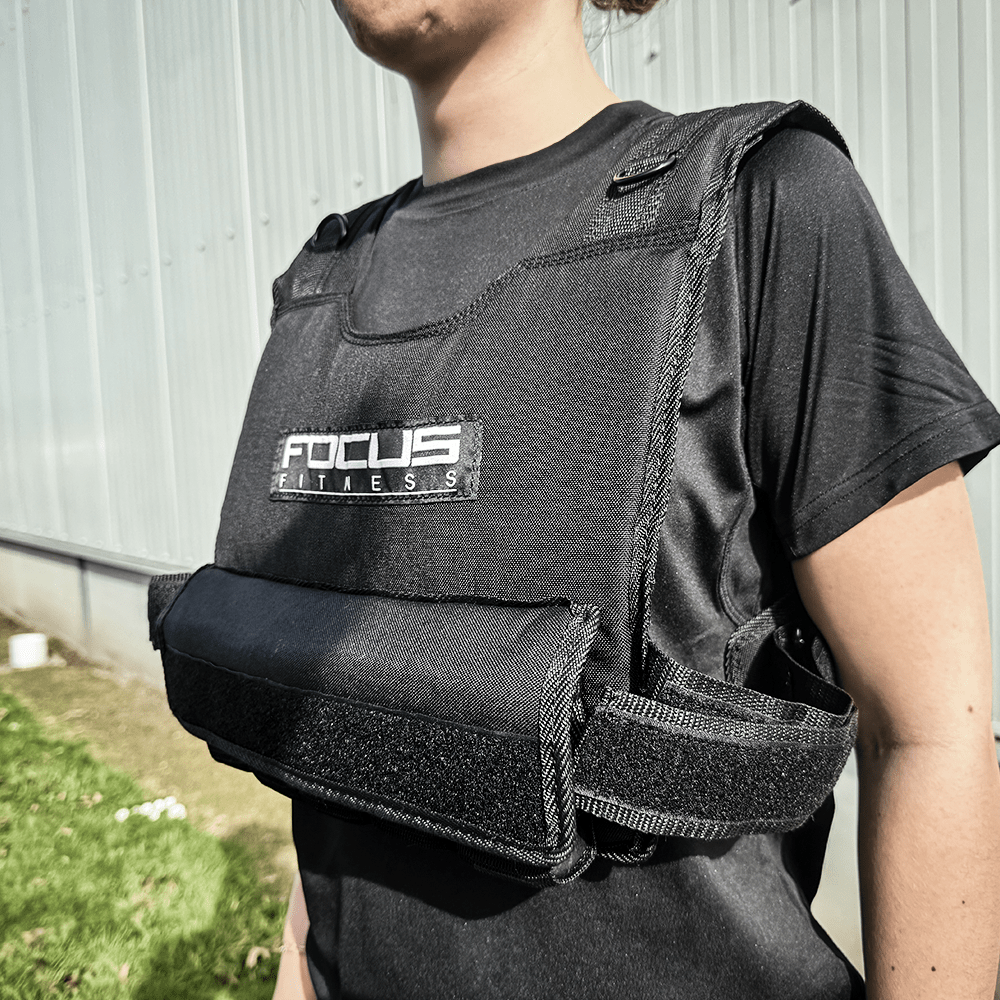 Personne portant un gilet lesté noir avec le logo Focus Fitness, debout près d'un mur gris.