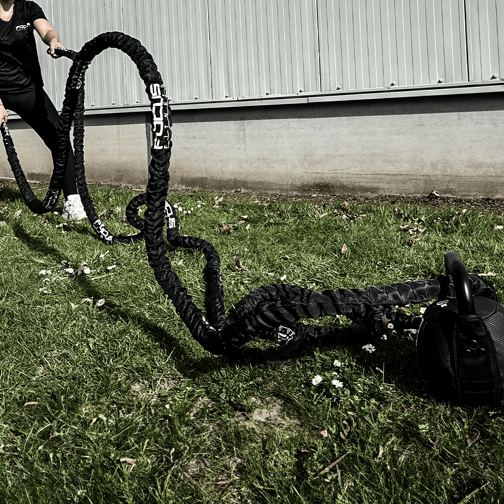 A person pulls black battle ropes on grass. A kettlebell rests in the background.