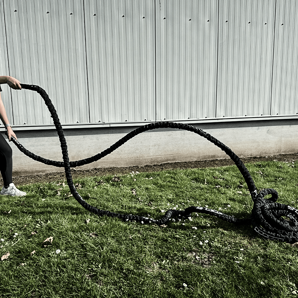 Person holding thick black battle ropes on a grassy field, near a white wall.