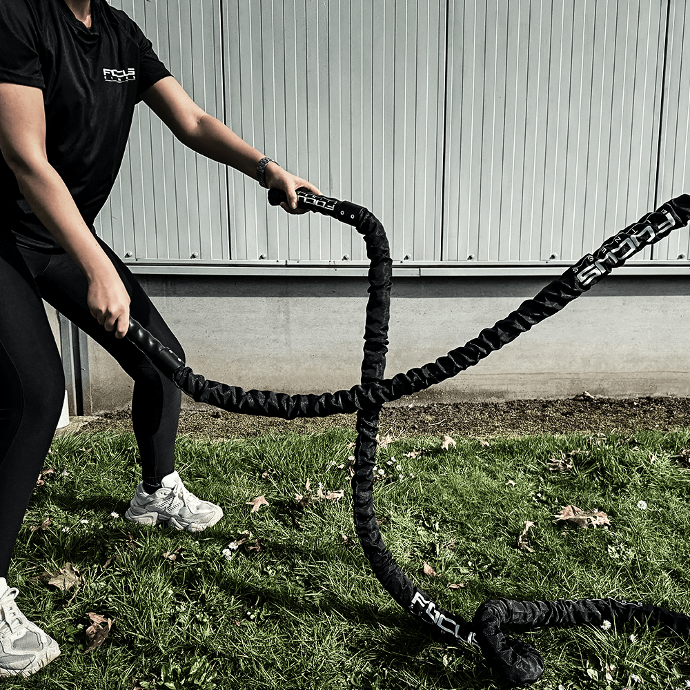 Two people training with black battle ropes on green grass. One is holding a rope handle.