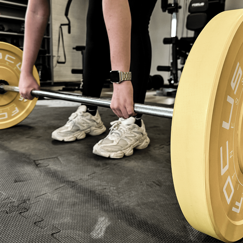 Personne soulevant une barre avec des poids jaunes dans une salle de sport. Ils portent des leggings noirs et des chaussures blanches.