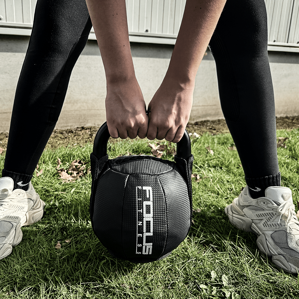 Person holding a black Focus Fitness kettlebell with both hands on green grass, legs apart.