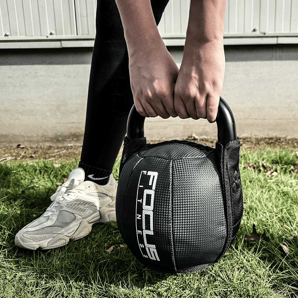 Person holding a black Focus Fitness kettlebell with both hands on green grass, legs apart.