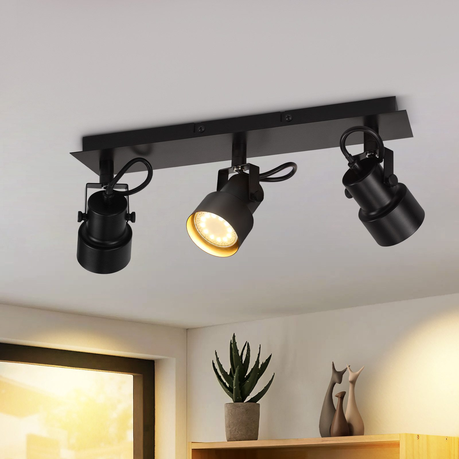 Trzy czarne oprawy reflektorów zamontowane na czarnym drążku. Tłem jest biały sufit i półka z dekoracjami.