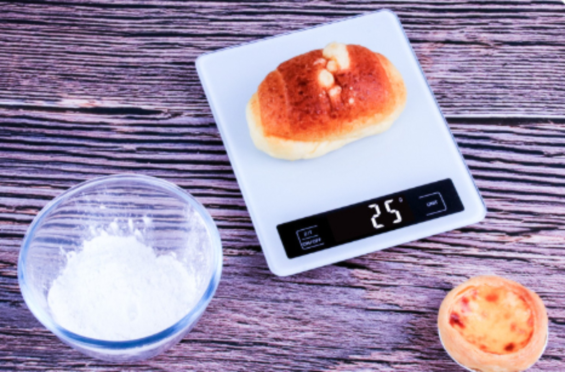 Ein Brötchen auf einer Digitalwaage mit 25 Gramm, neben einer Schüssel mit Mehl und einem kleinen Gebäck.