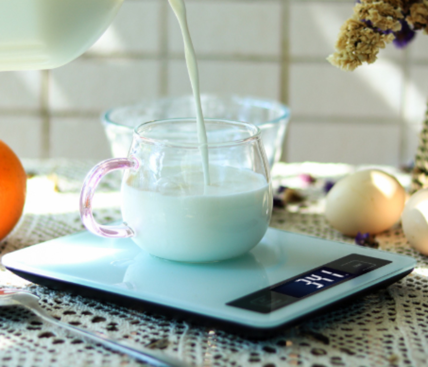 Milch wird in einen Glasbecher auf einer Waage gegossen, mit Ei und Blumen in der Nähe.
