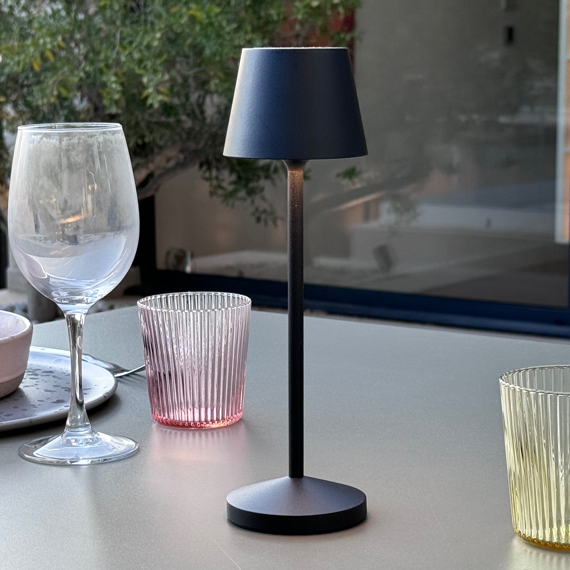 Schwarze Tischlampe mit schwarzem Schirm, neben Weinglas und rosa Glas.