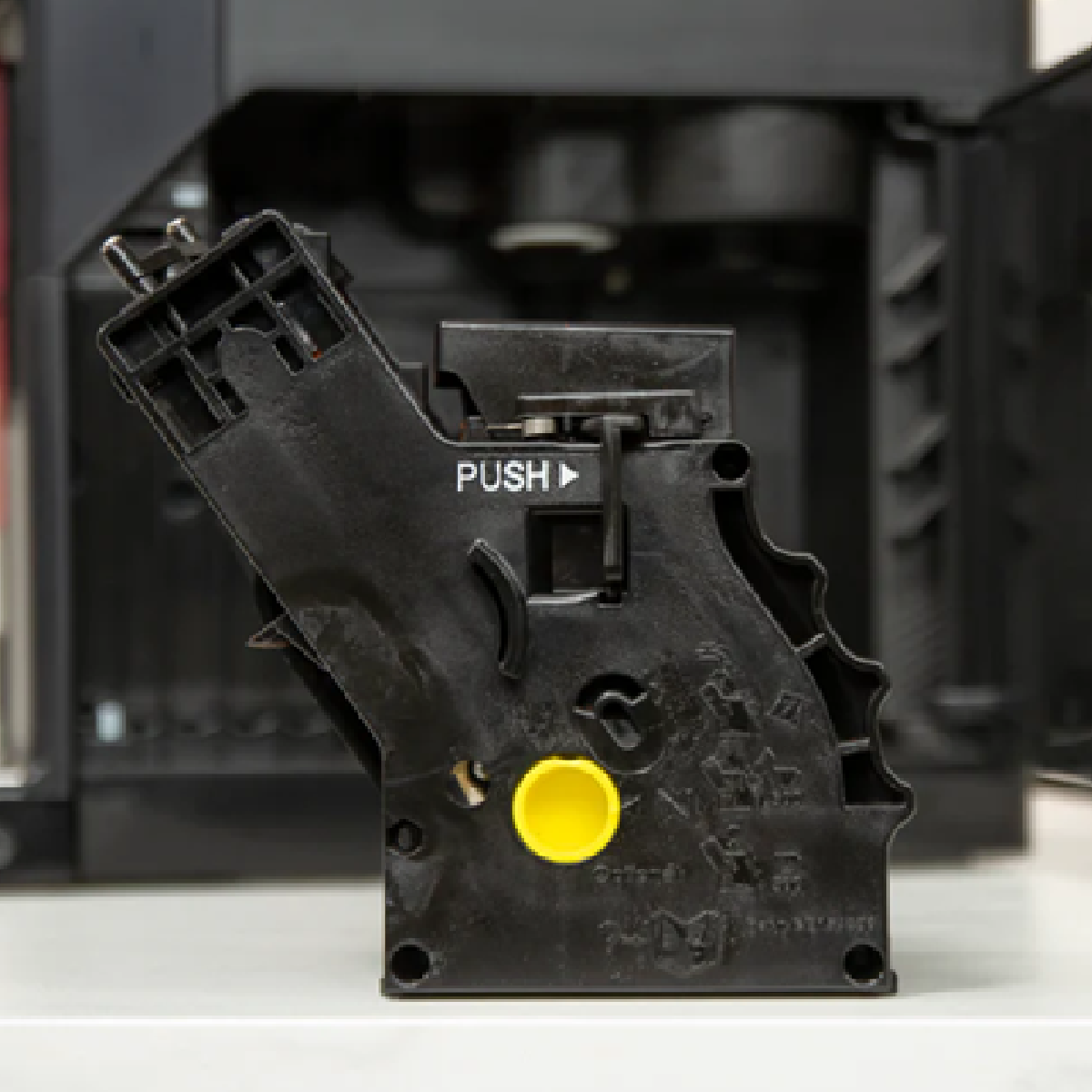 Czarny element ekspresu do kawy z żółtym przyciskiem, napisem "PUSH" i symbolem strzałki.