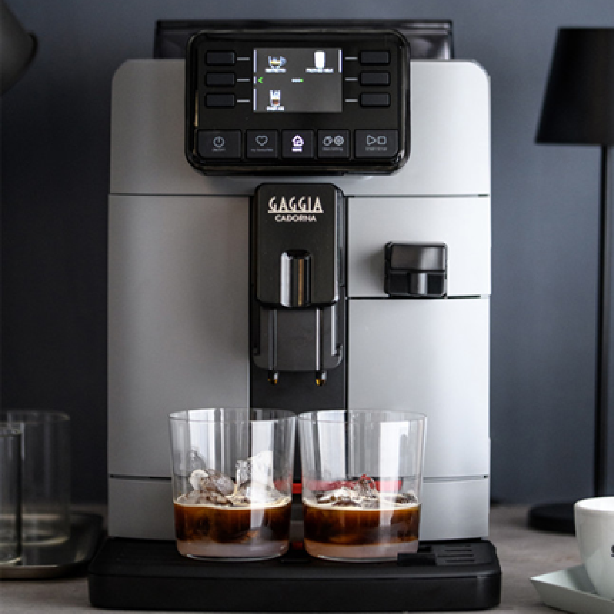 Een grijze koffiemachine met twee glazen koffie met ijs, op een zwart oppervlak.