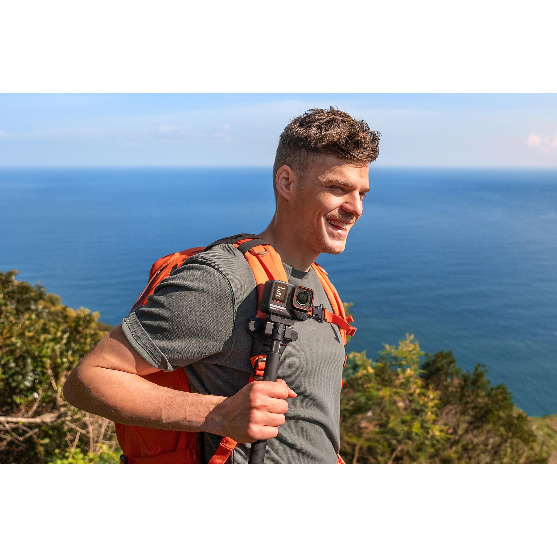 Mężczyzna z plecakiem trzymający aparat, uśmiechnięty, nad morzem.