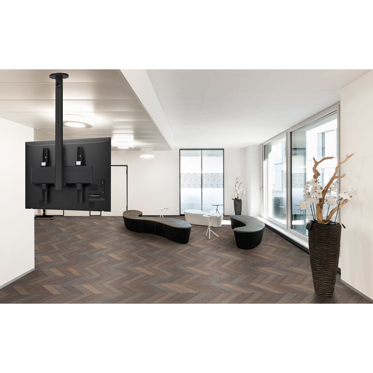 Großer Raum mit Fischgrät-Holzboden. Ein TV hängt von der Decke. Möbel umfassen schwarze Bänke.